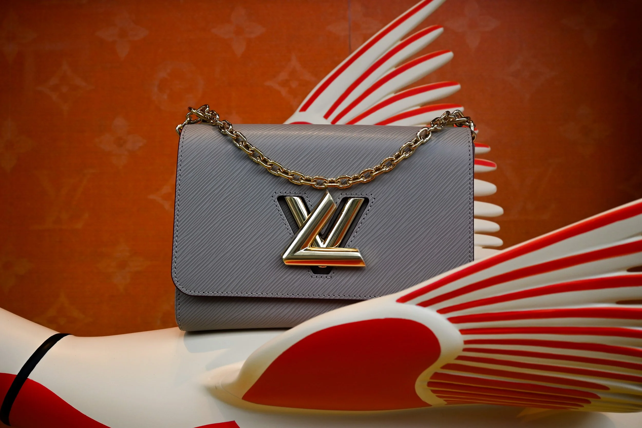 A gray designer handbag with a gold chain strap and a metallic gold logo on the front, displayed against a background of abstract red and white sculptures.