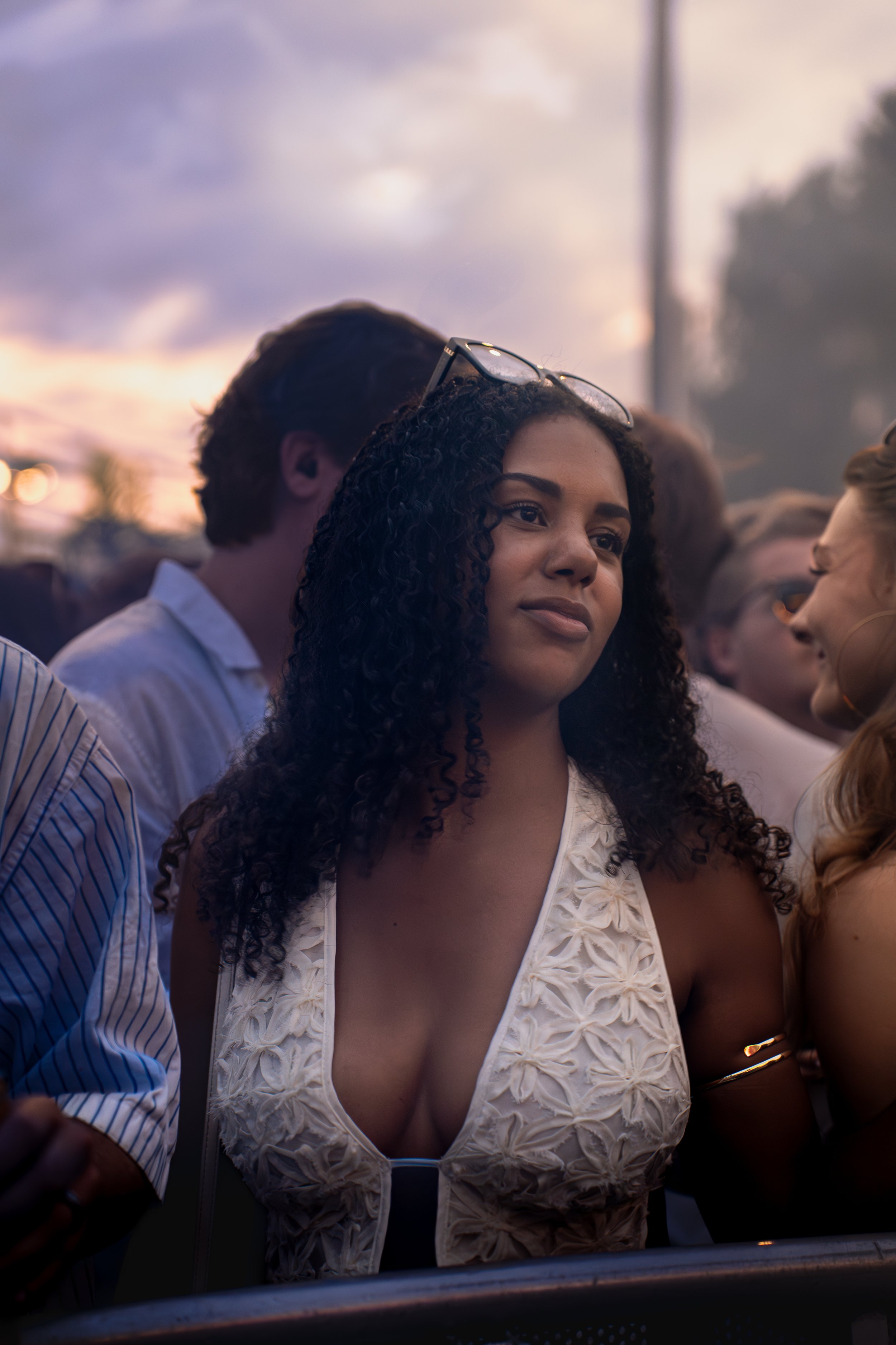 A woman with curly hair wearing glasses on her head and a white, detailed dress, standing outdoors during sunset, surrounded by other people, at what appears to be a social event or gathering.