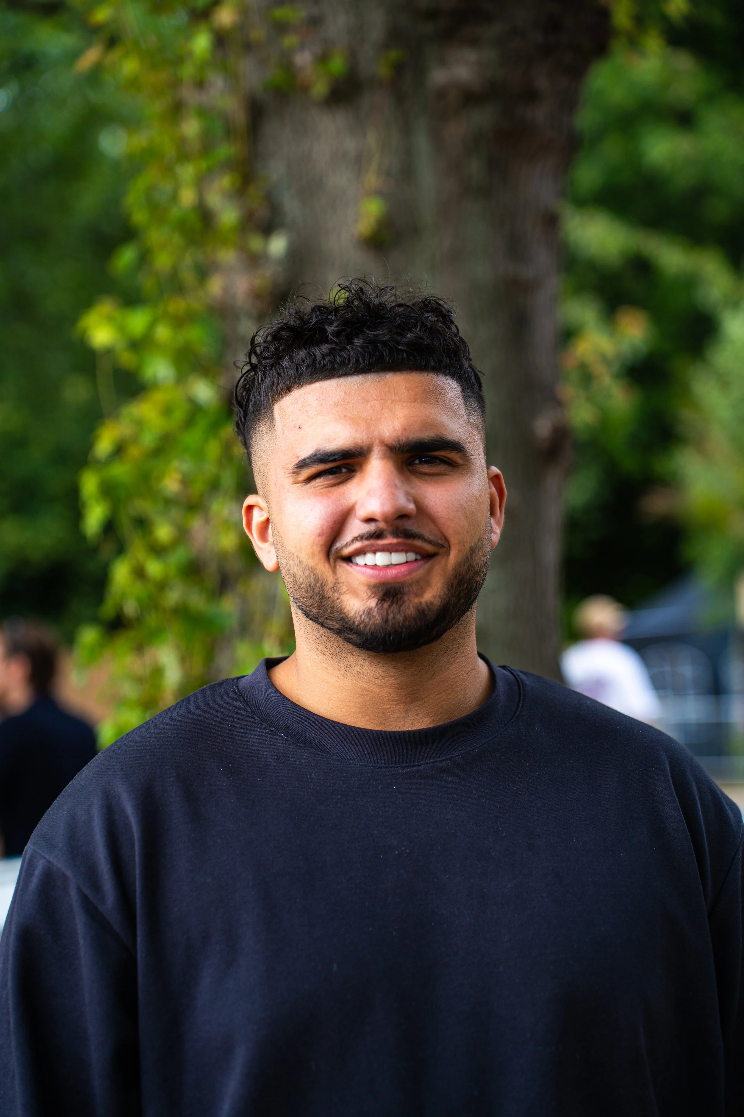 A young man with short, curly black hair and a beard, smiling outdoors in front of a tree and greenery.