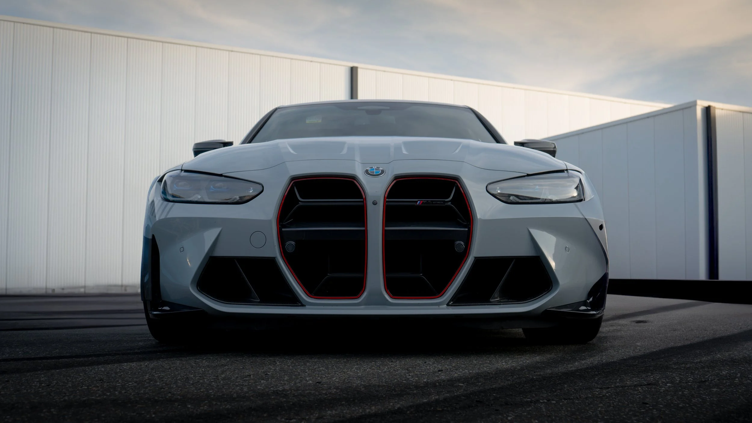 Front view of a white BMW M4 sports car with black and red accents parked on asphalt, with industrial buildings in the background and a cloudy sky above.
