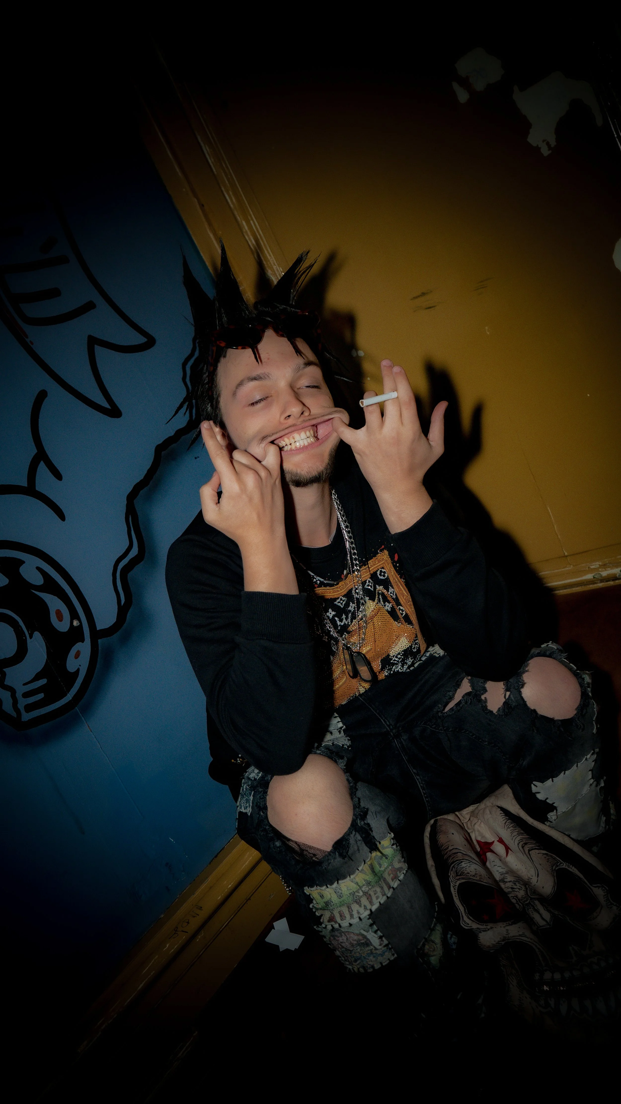 A young man with spiked hair, wearing distressed ripped jeans and graphic patches, smiling with one eye closed, holding a cigarette in his right hand, sitting against a colorful wall with cartoon artwork.