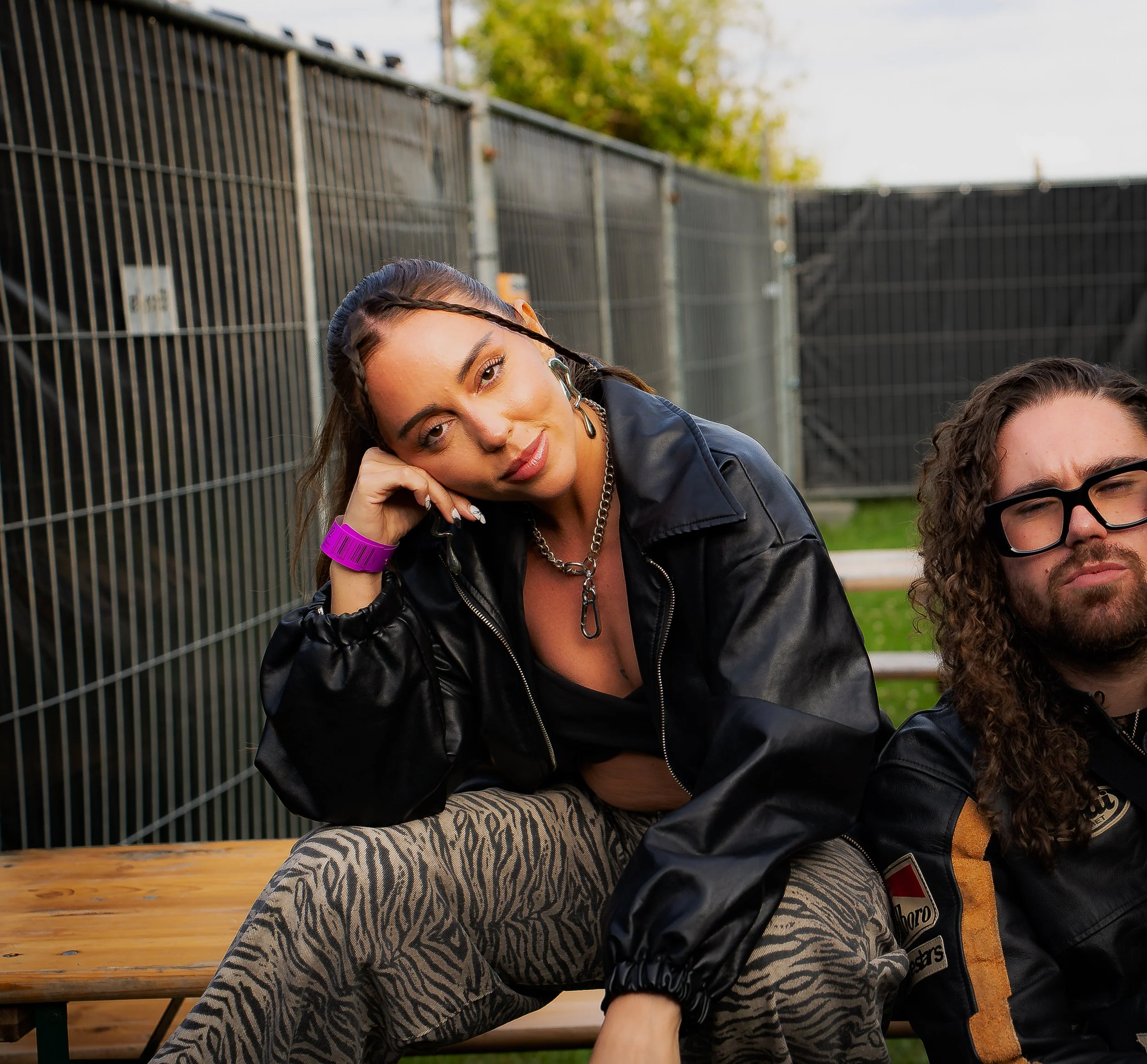 A young woman with braided hair and a woman with curly hair sitting on a wooden bench outdoors, with a chain-link fence and trees in the background.