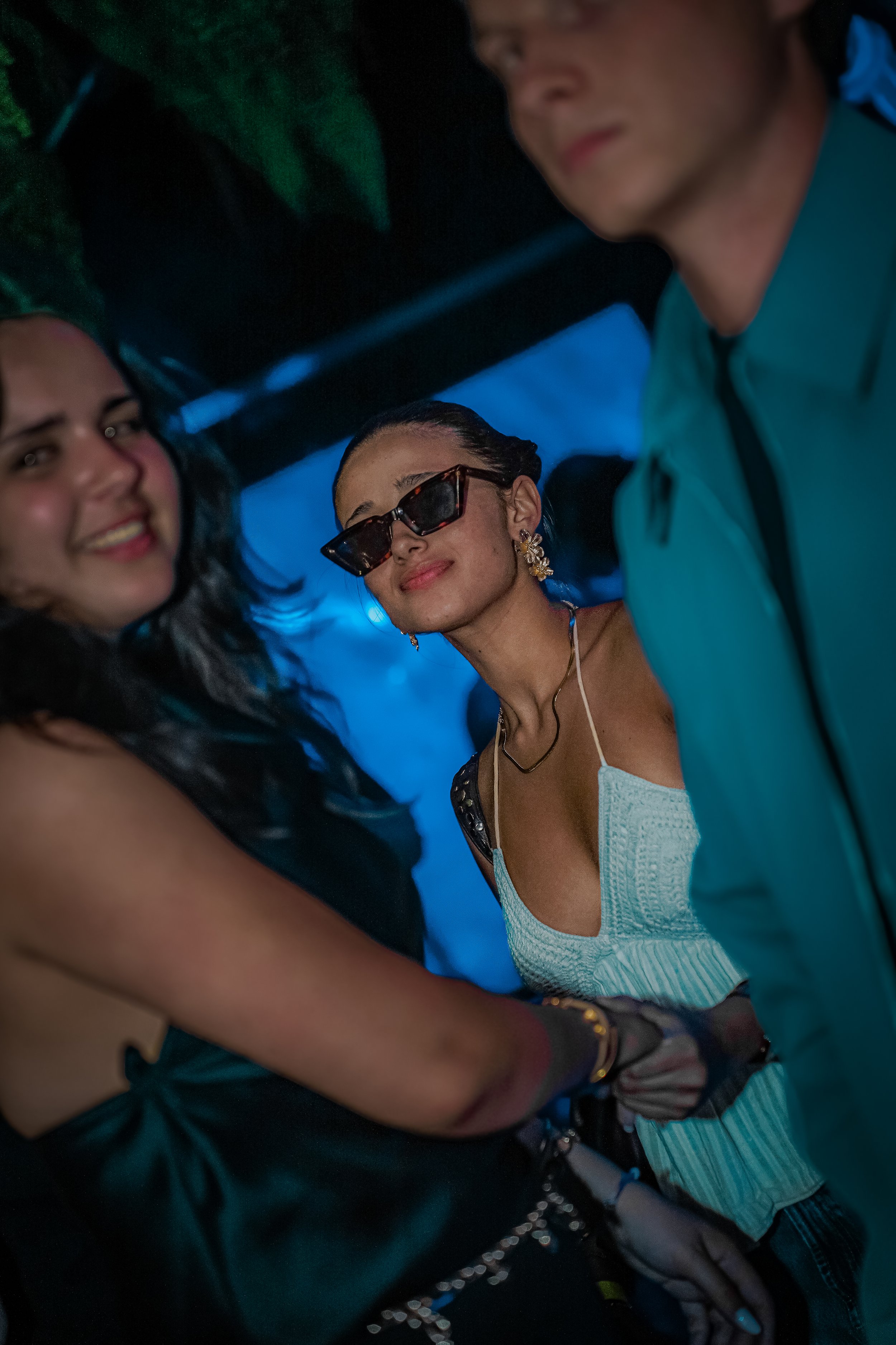 Three people are in a dark, lively setting. The woman in the center is wearing sunglasses, earrings, and a white top, smiling confidently. The woman on the left has black hair, is smiling, and wearing a black outfit. The man on the right is wearing a