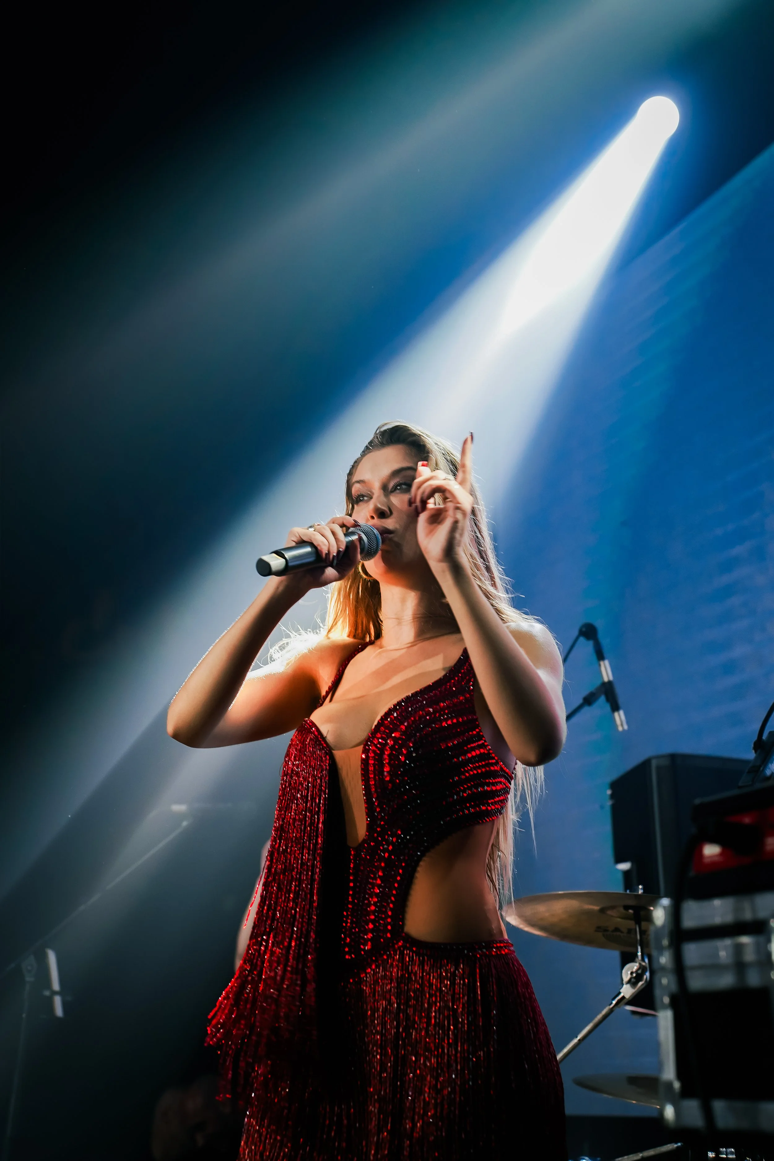 A female singer performs on stage, holding a microphone and pointing upward with one finger. She is wearing a red, beaded, cut-out dress and is illuminated by a bright spotlight.
