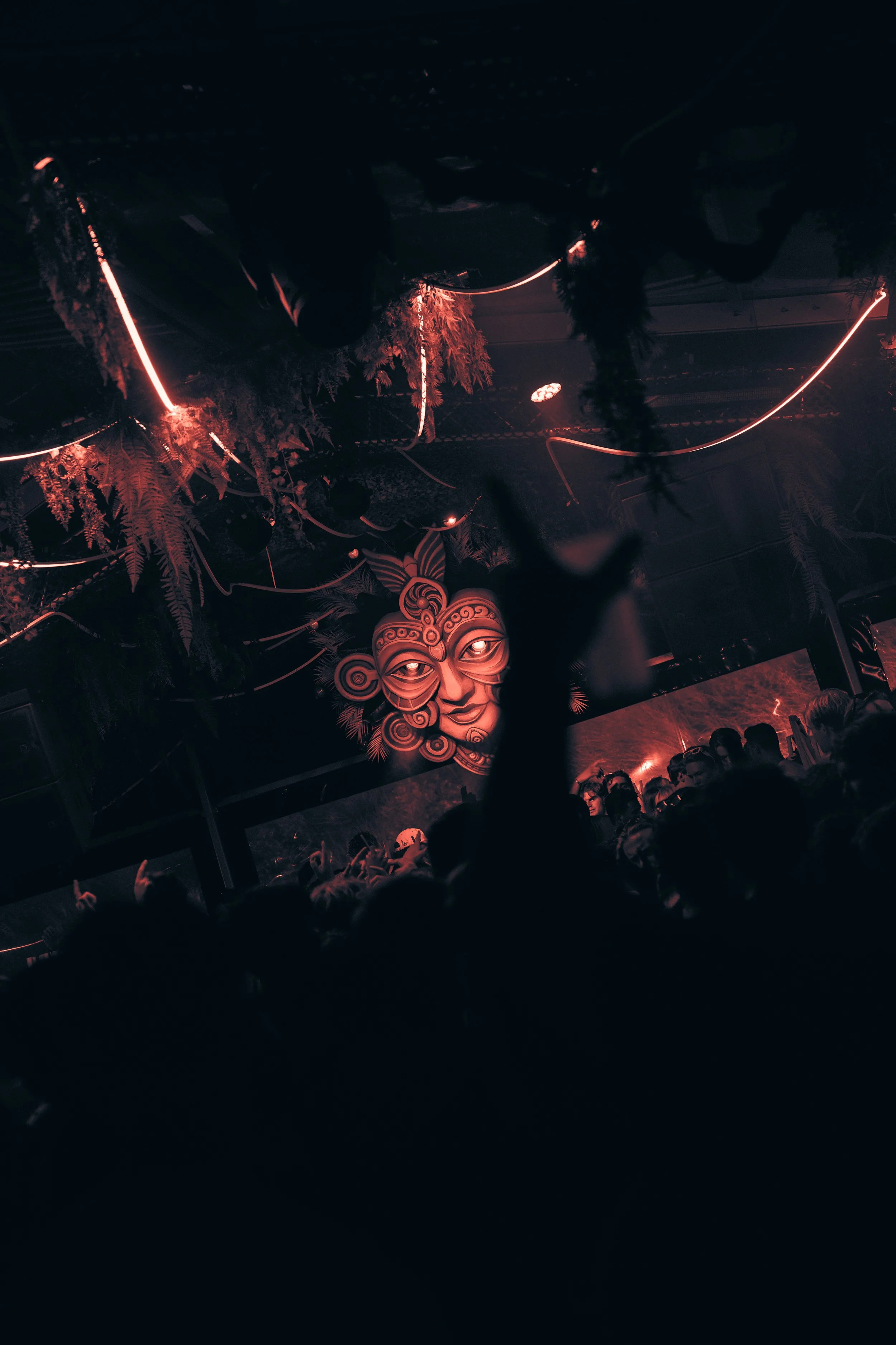 Dark indoor concert scene with a large tribal face mask decoration on the wall, green hanging foliage, and red lighting; crowd of people in the foreground with one arm raised.