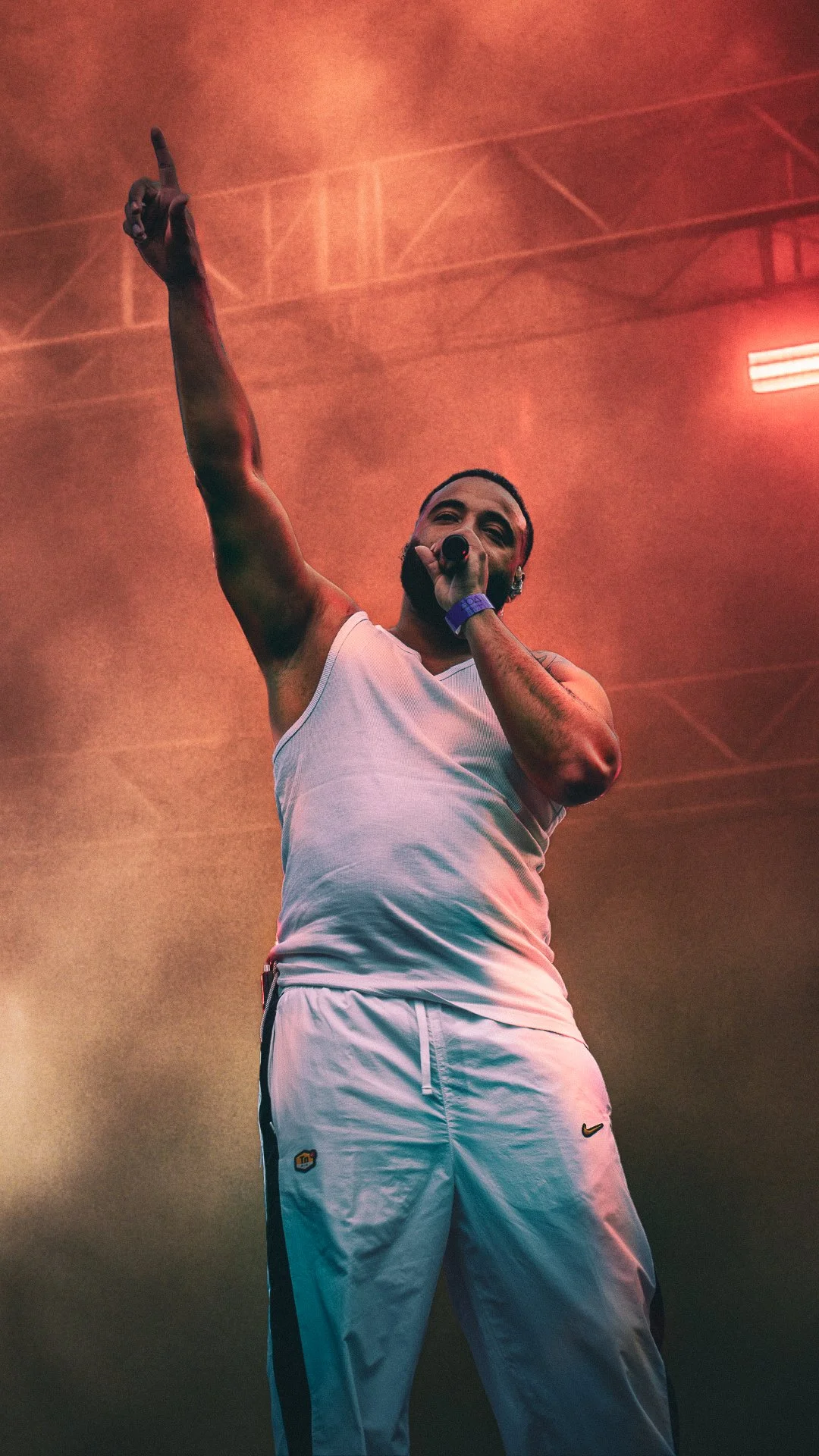 A male performer on stage wearing a white tank top and white pants, holding a microphone, raising his right arm with his index finger pointing up, under red stage lighting.
