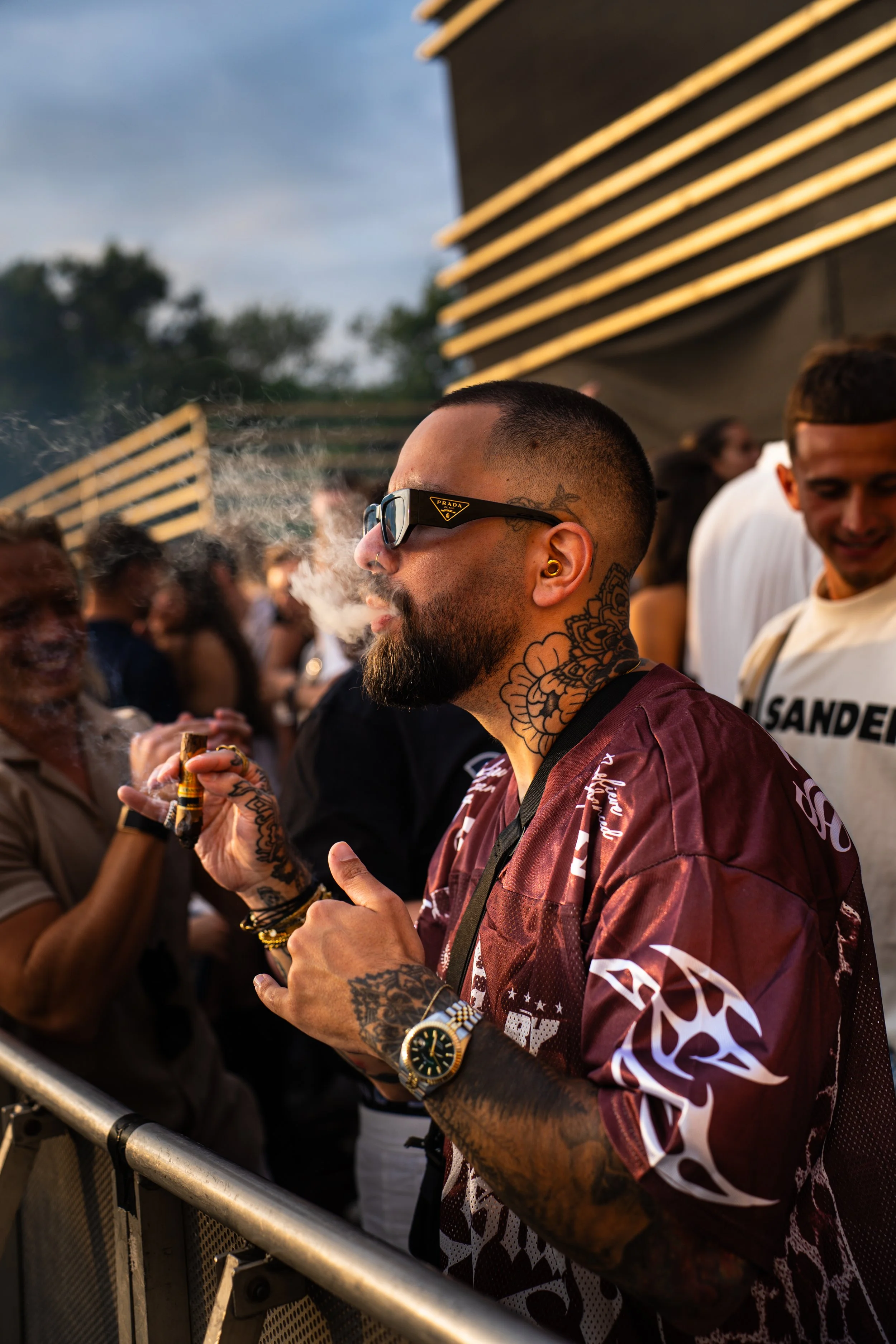 Man with sunglasses, tattoos, and a beard smoking a cigar at an outdoor event with a crowd.