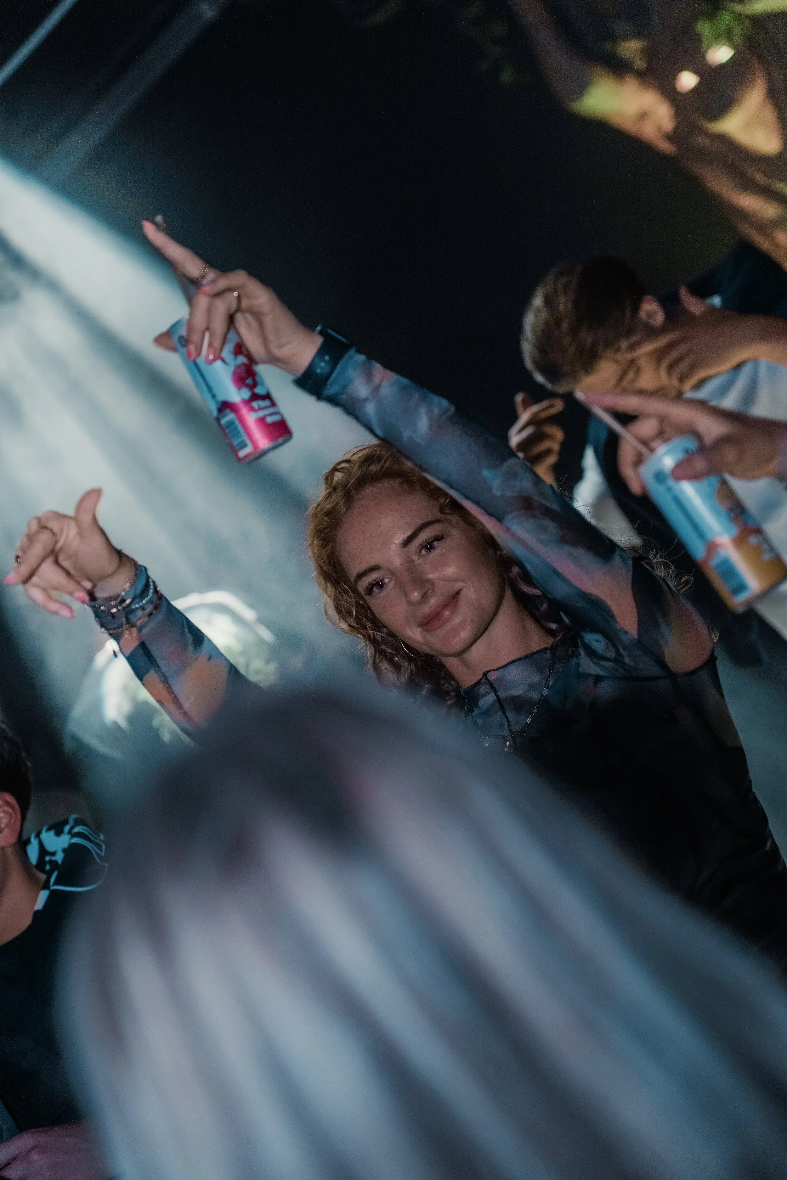 Person with curly hair and a dark top dancing at a party with others around, holding a can of Red Bull.