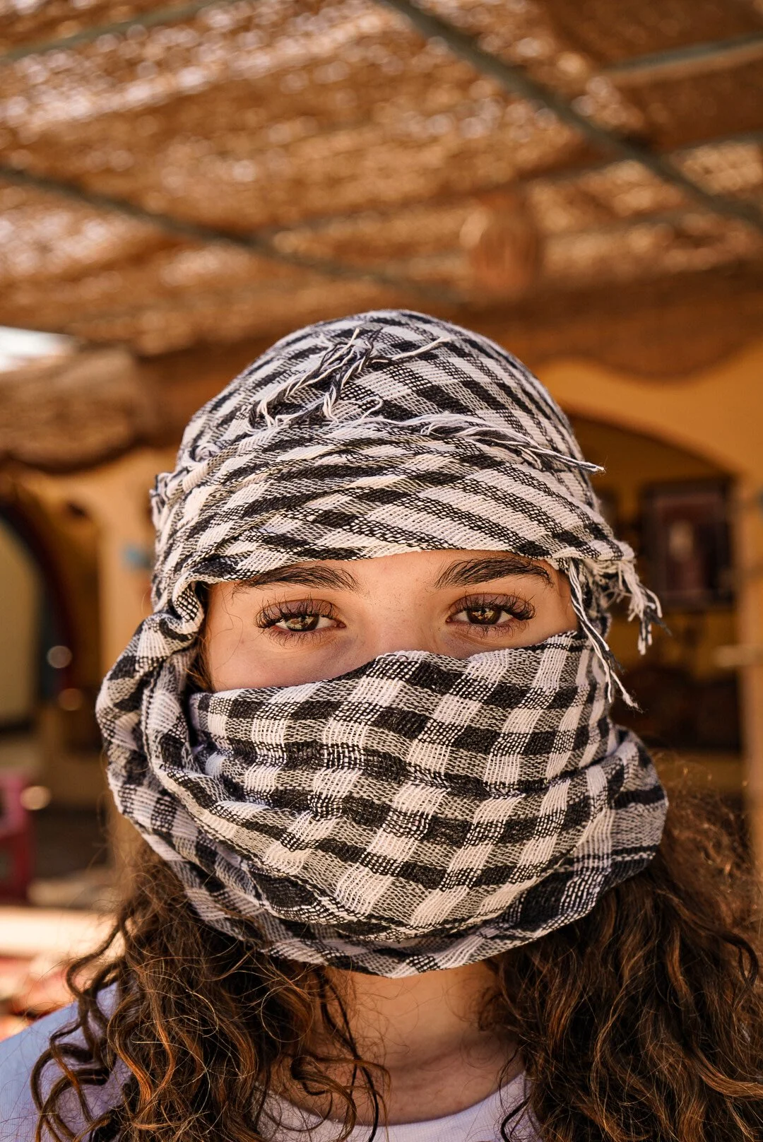 A person with curly hair wearing a black and white checkered headscarf covering their face and head, with only their eyes visible, inside a building with a decorated ceiling.