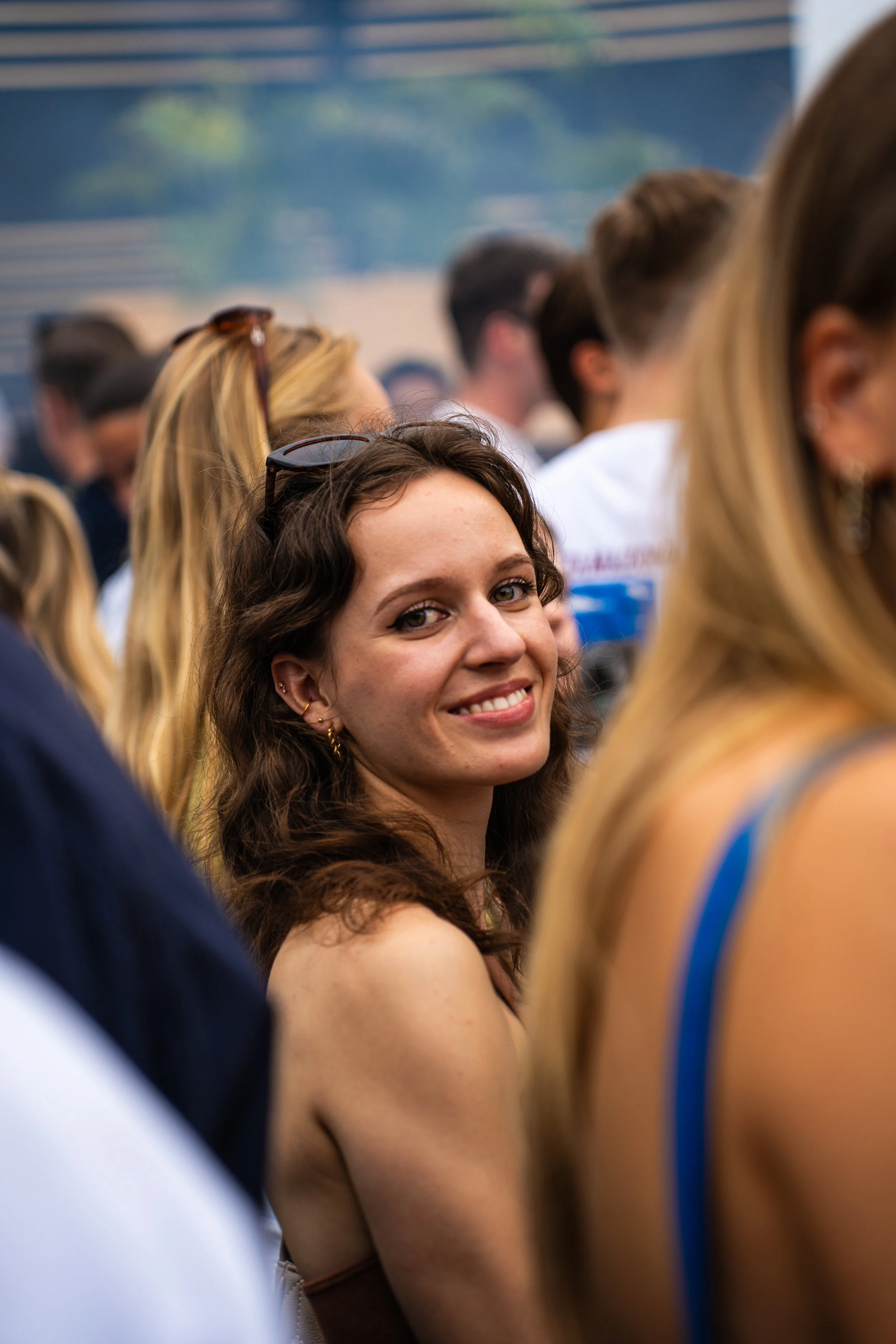 A young woman with wavy brown hair and earrings smiling at the camera among a crowd of people.