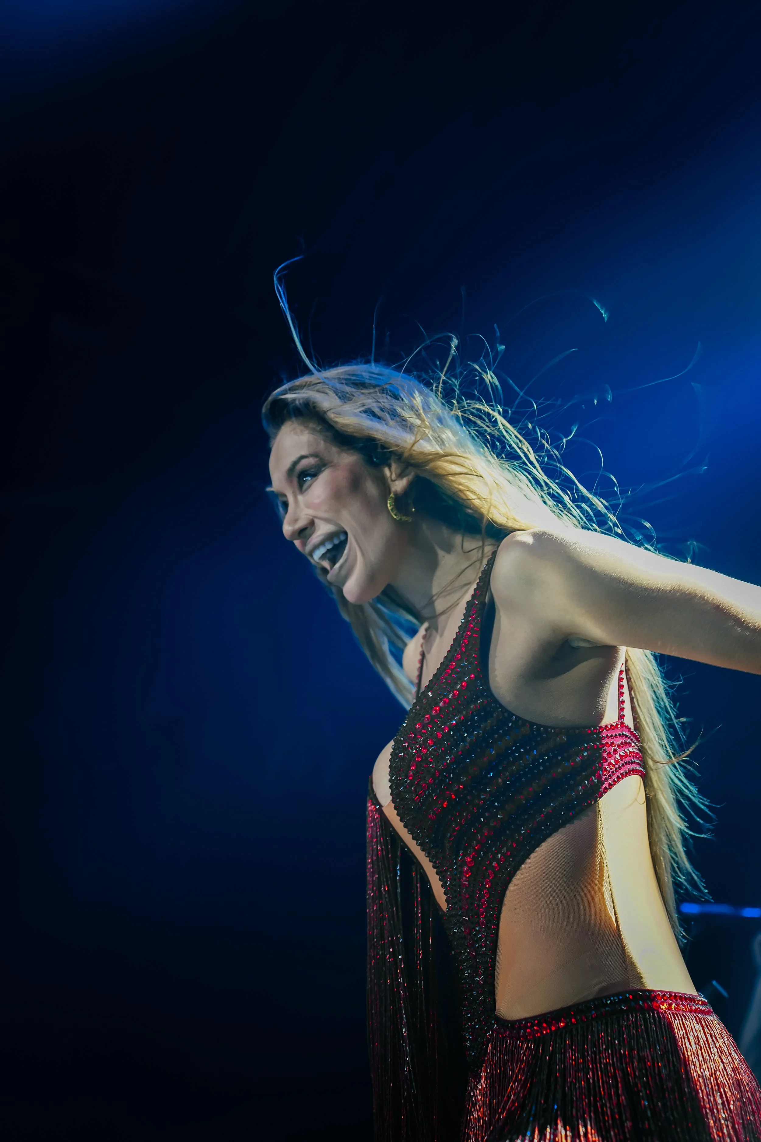 A woman with long hair smiling, wearing a black and red sequined dress, on stage with blue lighting.