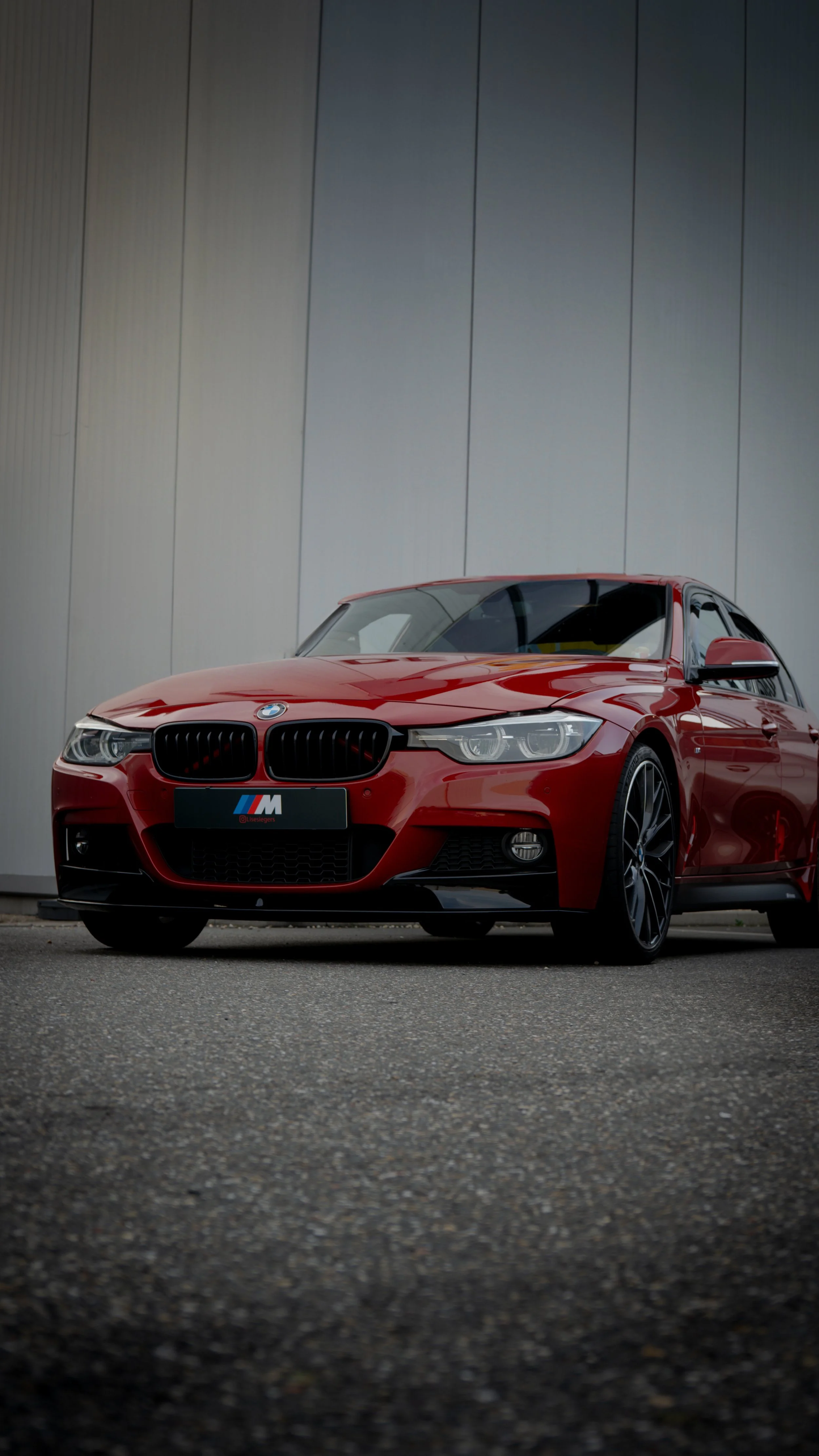 Red BMW M series car parked on a textured pavement in front of a modern metallic wall.