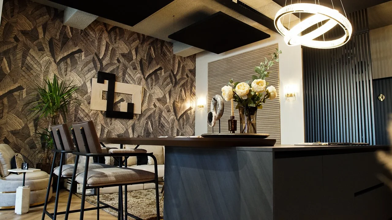 Modern living room with patterned wallpaper, black and white geometric wall art, beige sofa, black chairs, a plant, and a black kitchen island with white flowers in a vase.