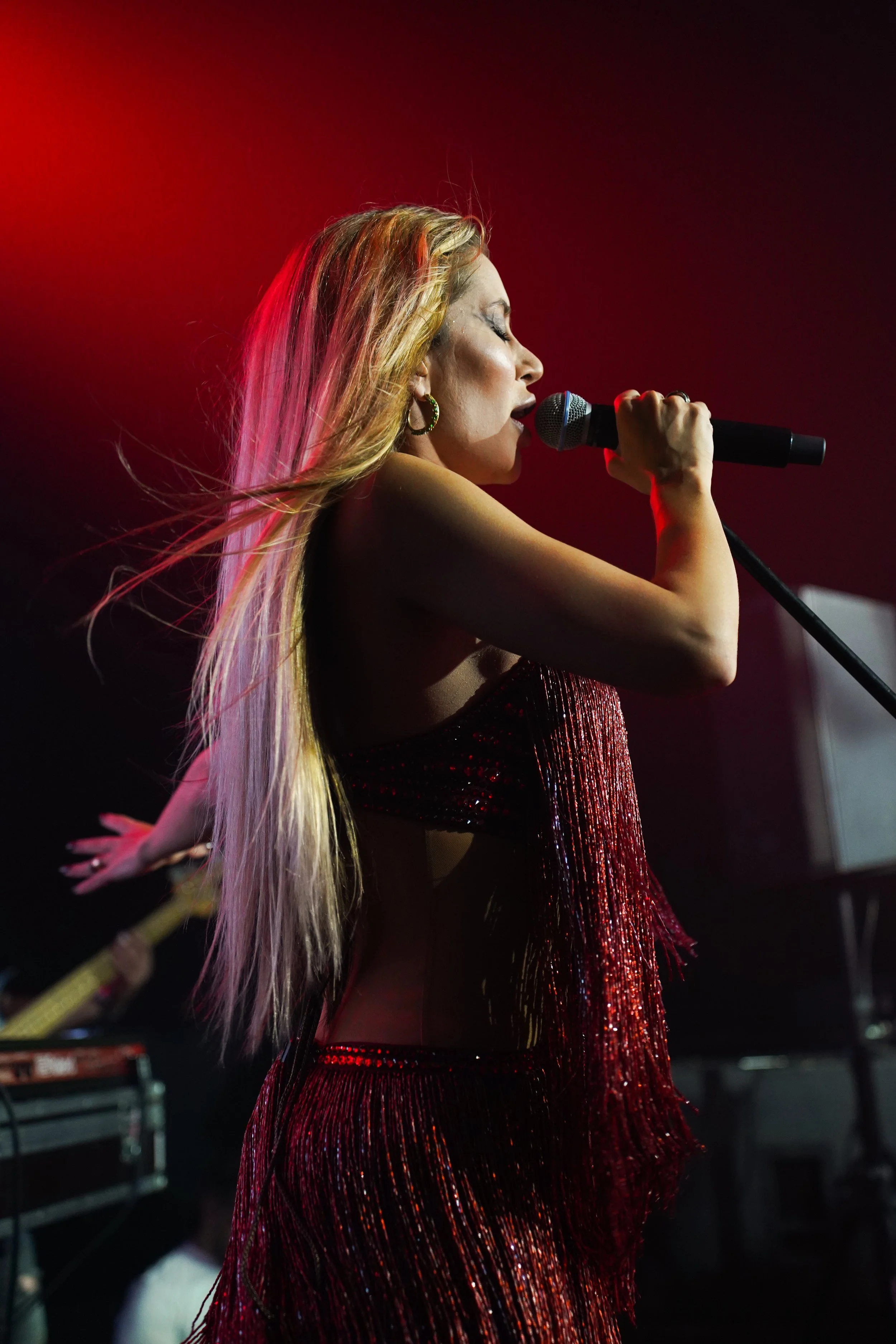 A female singer with long blonde hair performing on stage, holding a microphone with her eyes closed, wearing a sparkling red dress with fringe details, against a dark background with red lighting.