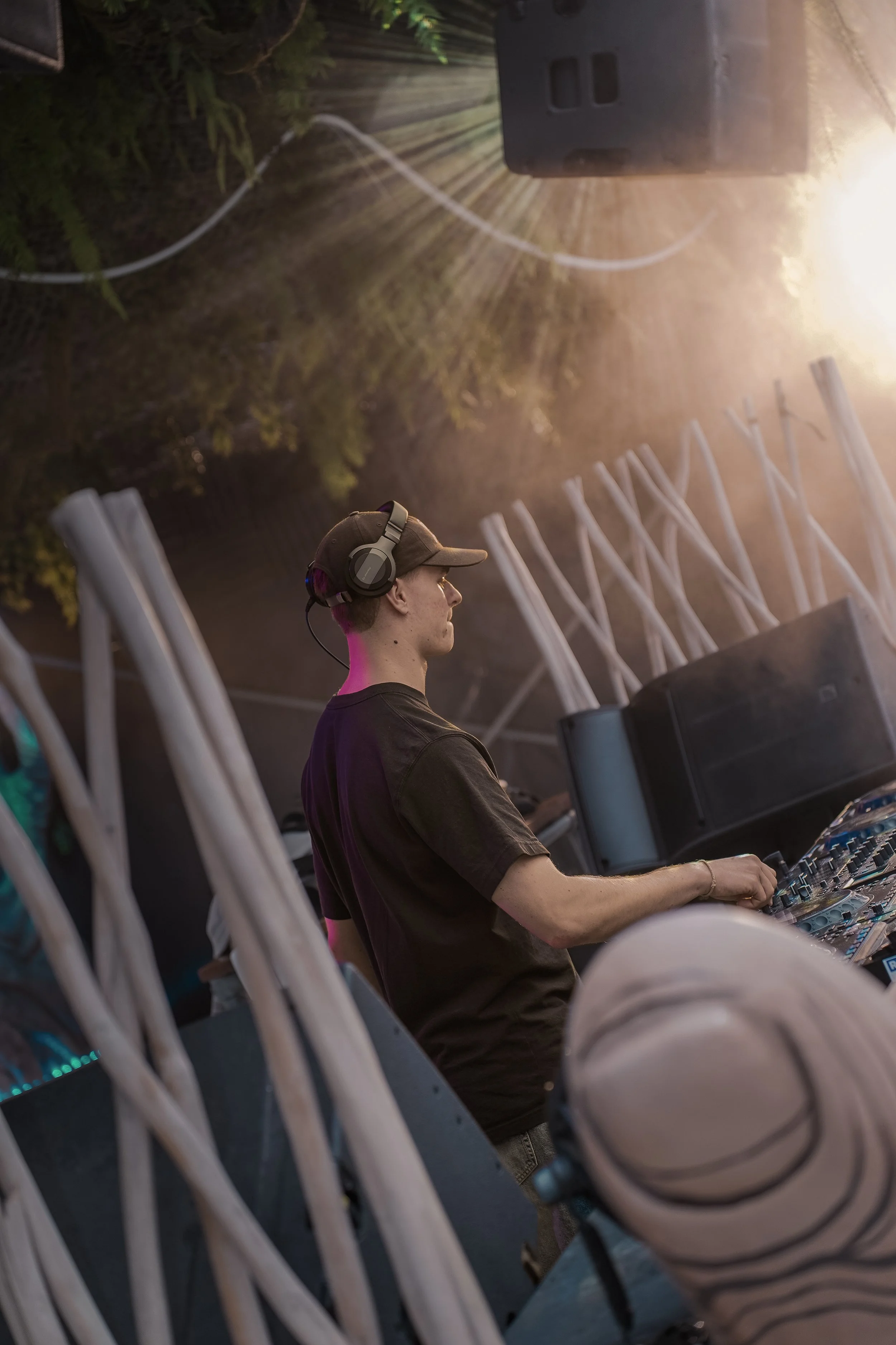 A DJ wearing a cap and headphones performs at an outdoor music event during sunset, with trees and stage equipment in the background.