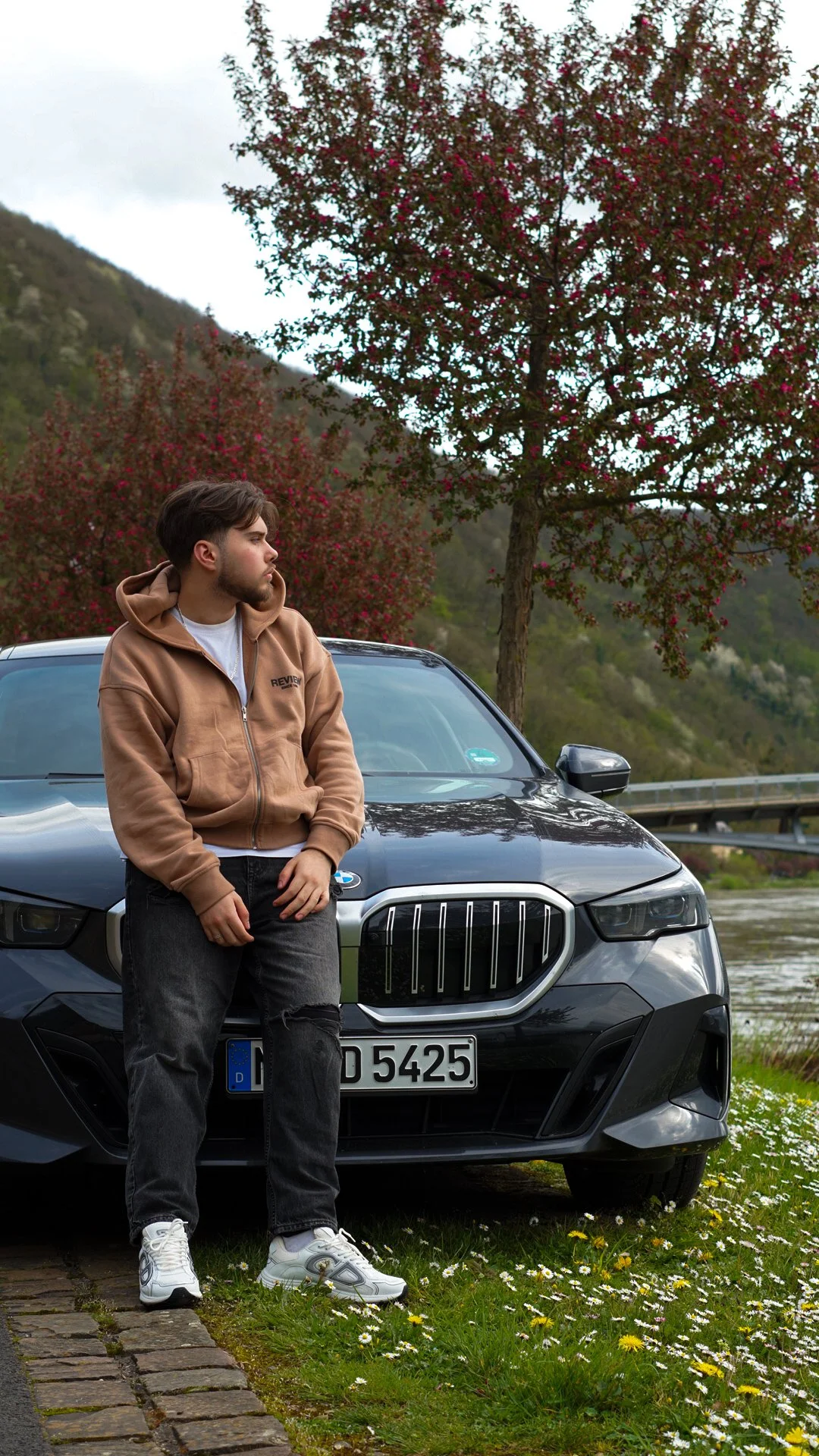 A young man with light skin and dark hair leans against the front of a black BMW parked by a river with a scenic mountain background. He is wearing a tan hoodie, black ripped jeans, and white sneakers, and is looking off to the side. Behind him, there are trees with pink blossoms along the riverbank.