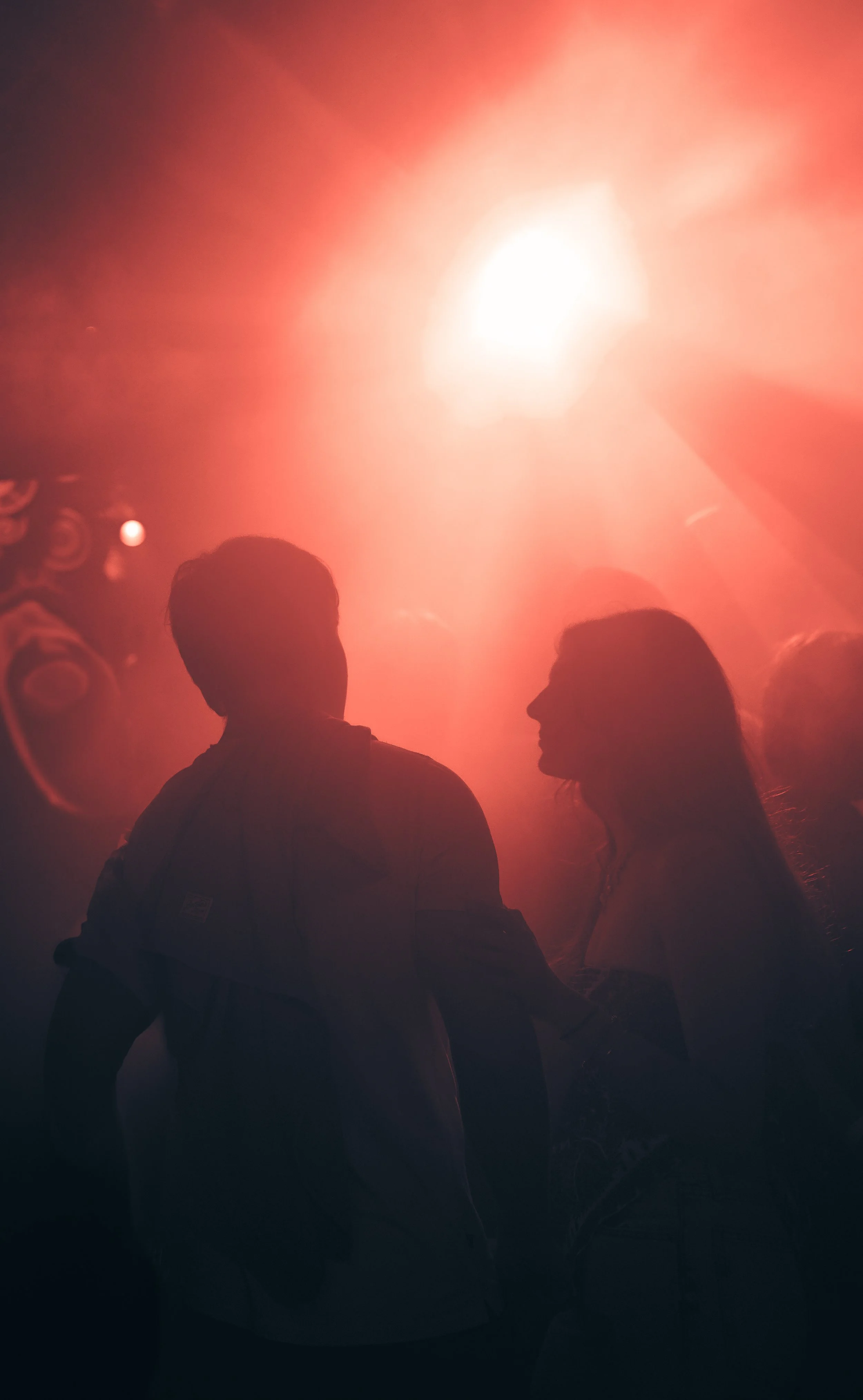 Silhouettes of two people under bright red and orange lighting at a crowded party or concert.