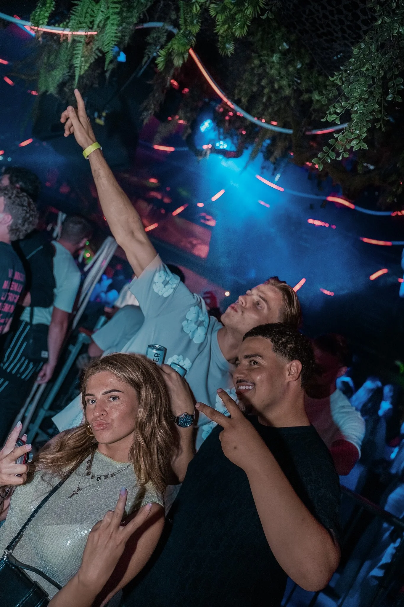 Three friends at a party, posing for a photo with peace signs. Two men and a woman, with colorful lighting and a dance floor in the background.