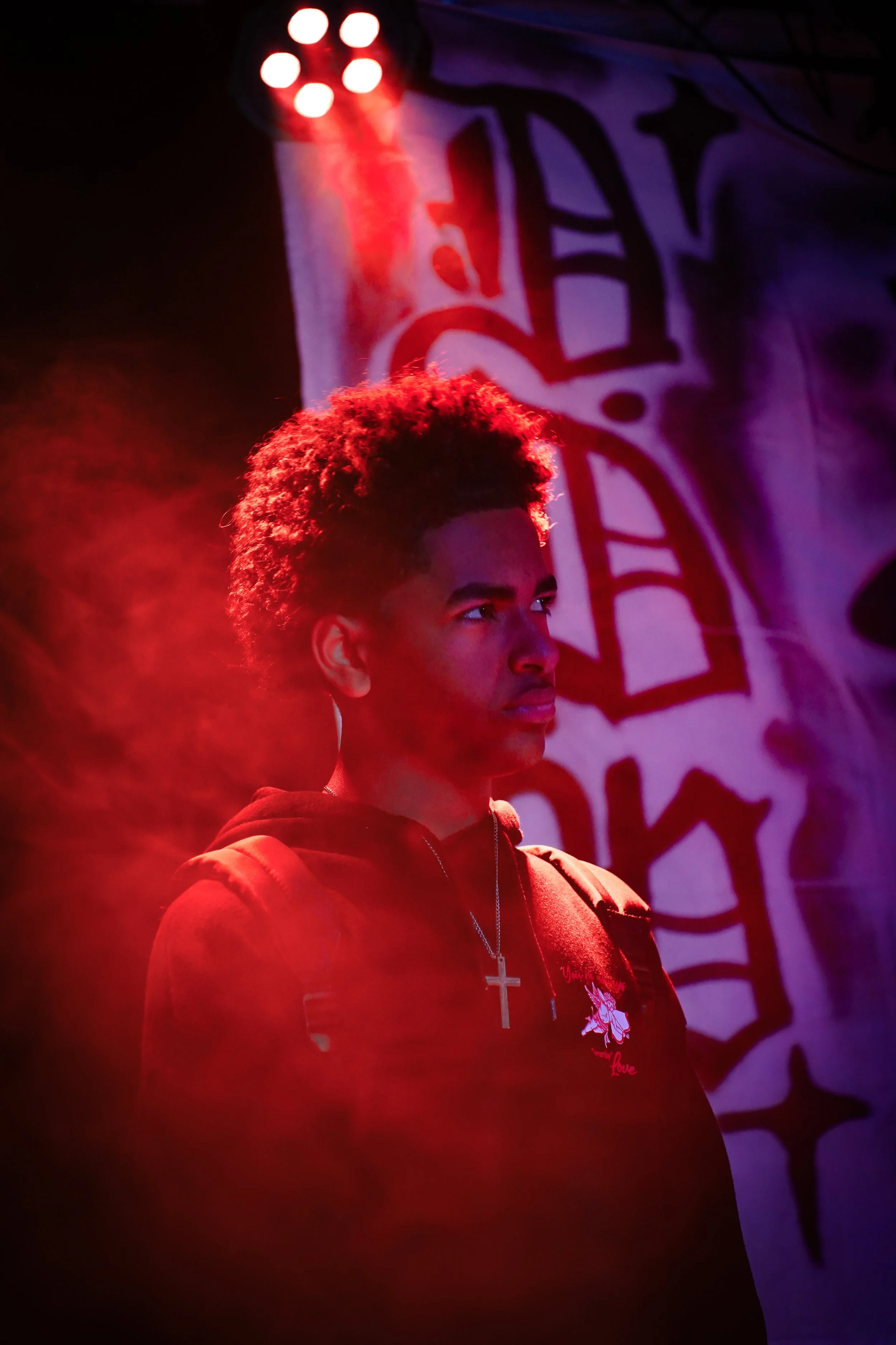 A young man with curly hair standing in dramatic red and blue lighting, with a graffiti-style mural in the background.