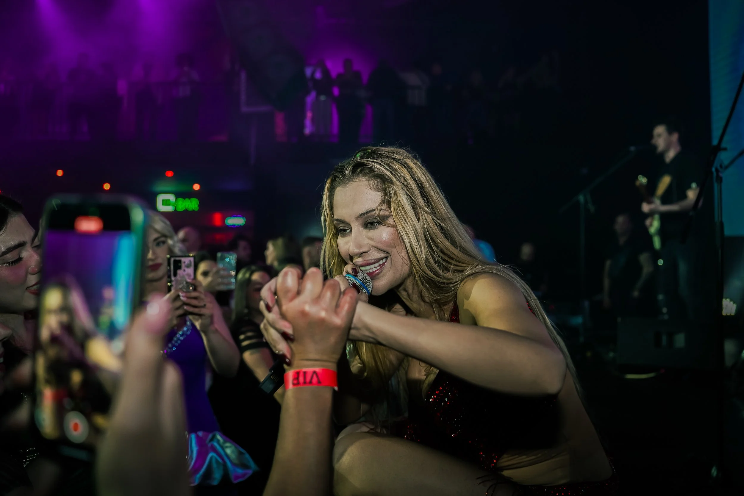 A female singer performing on stage at a concert, engaging with the audience and smiling while singing into a microphone, with fans taking photos around her and a guitarist in the background.