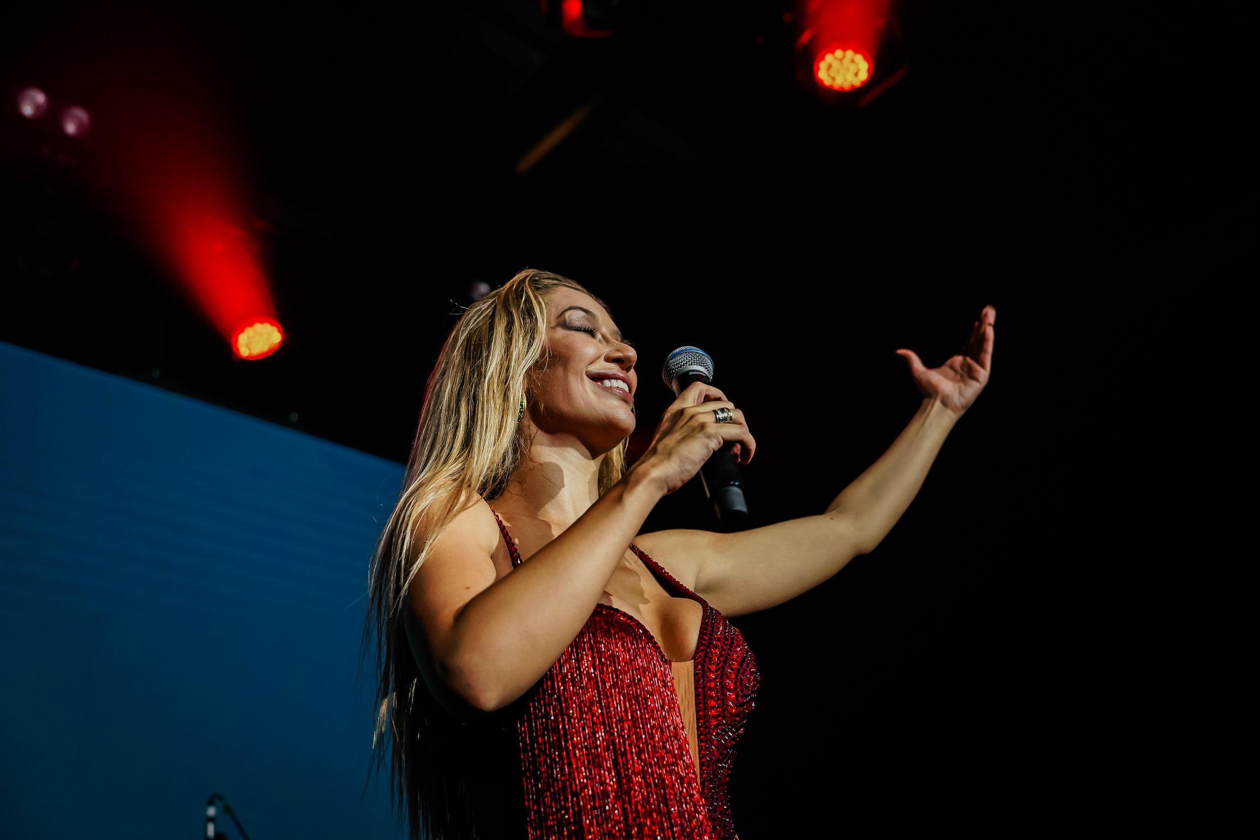 Woman in a red dress singing into a microphone on stage with red and blue lighting.