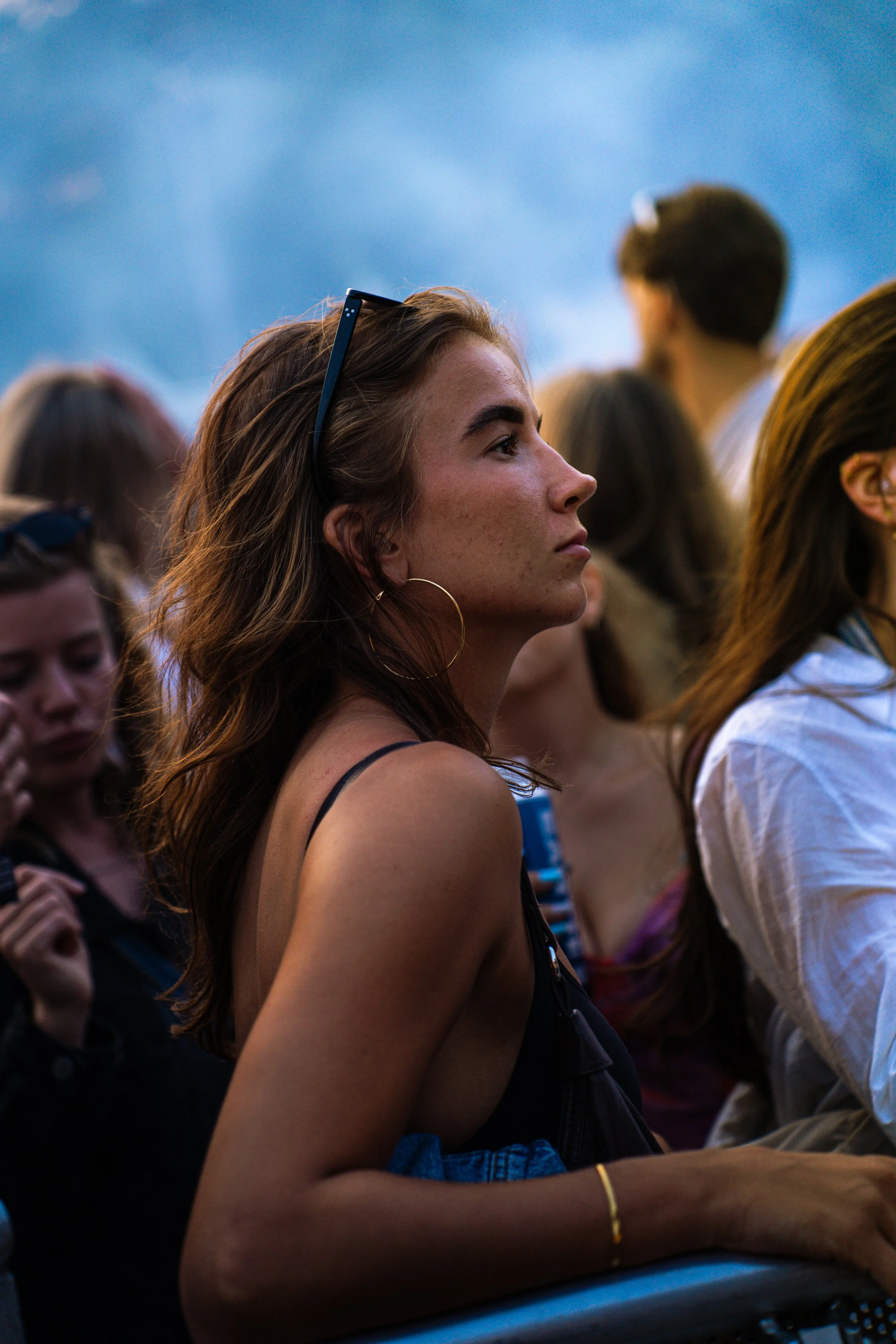 A young woman with long brown hair wearing hoop earrings and a sleeveless top, standing in a crowd outdoors during dusk, watching attentively.