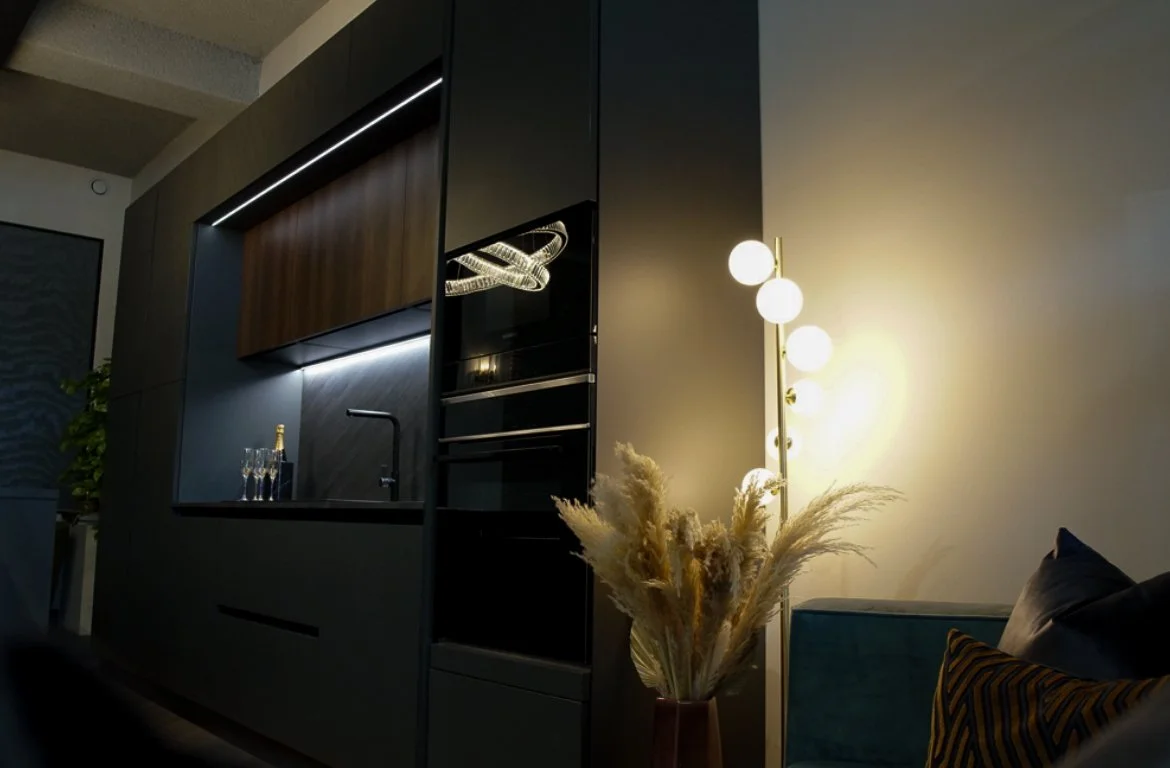 Modern kitchen and living room with a lit floor lamp, pampas grass in a vase, a dark sofa with pillows, and built-in oven in the dark cabinetry.