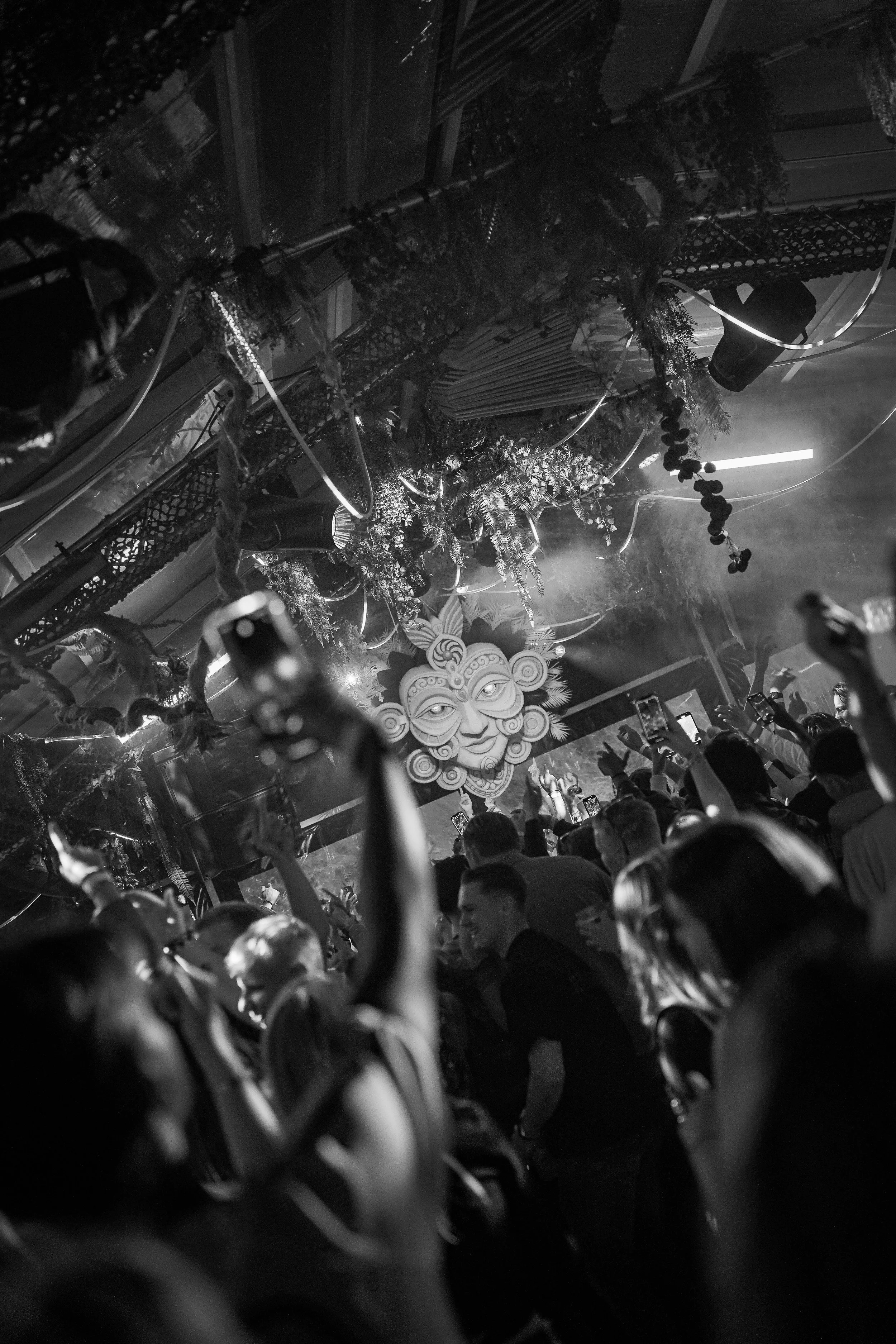 A black and white photo of a crowded dance floor at a nightclub with people taking photos and videos. There is a large, decorative face mask or sculpture on the stage, and the ceiling is decorated with hanging plants and decorations.