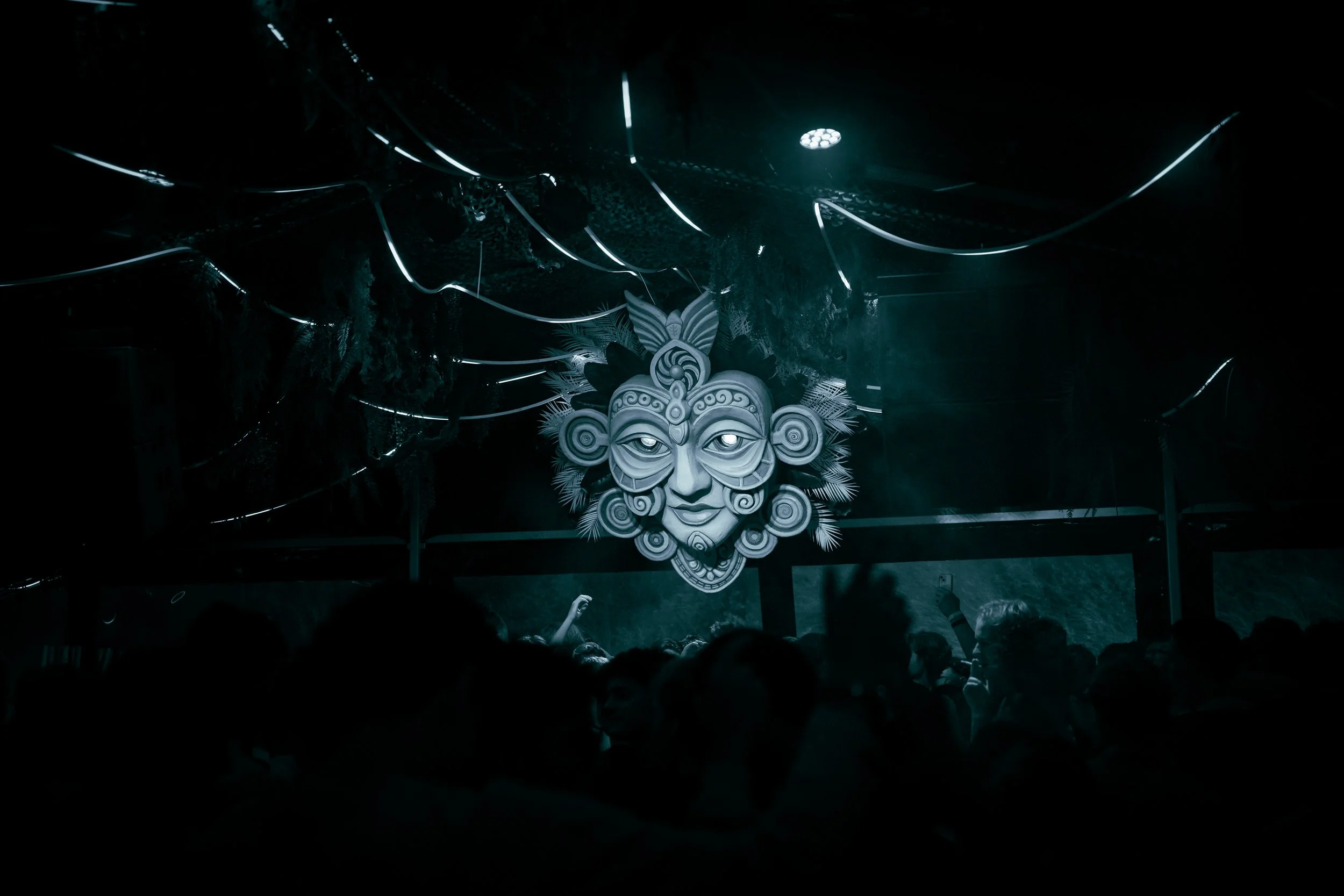 A crowd at a concert or festival with a large illuminated decorative mask or face on a wall, featuring a face with intricate patterns, feathers, and a headdress, under dark lighting.