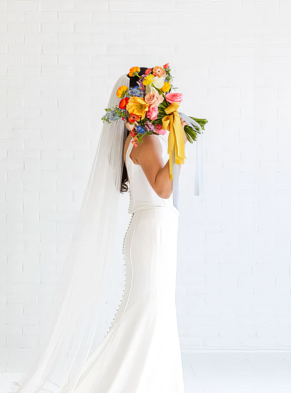 Woman in a white wedding dress holding a large colorful bouquet in front of her face, standing against a white brick wall.