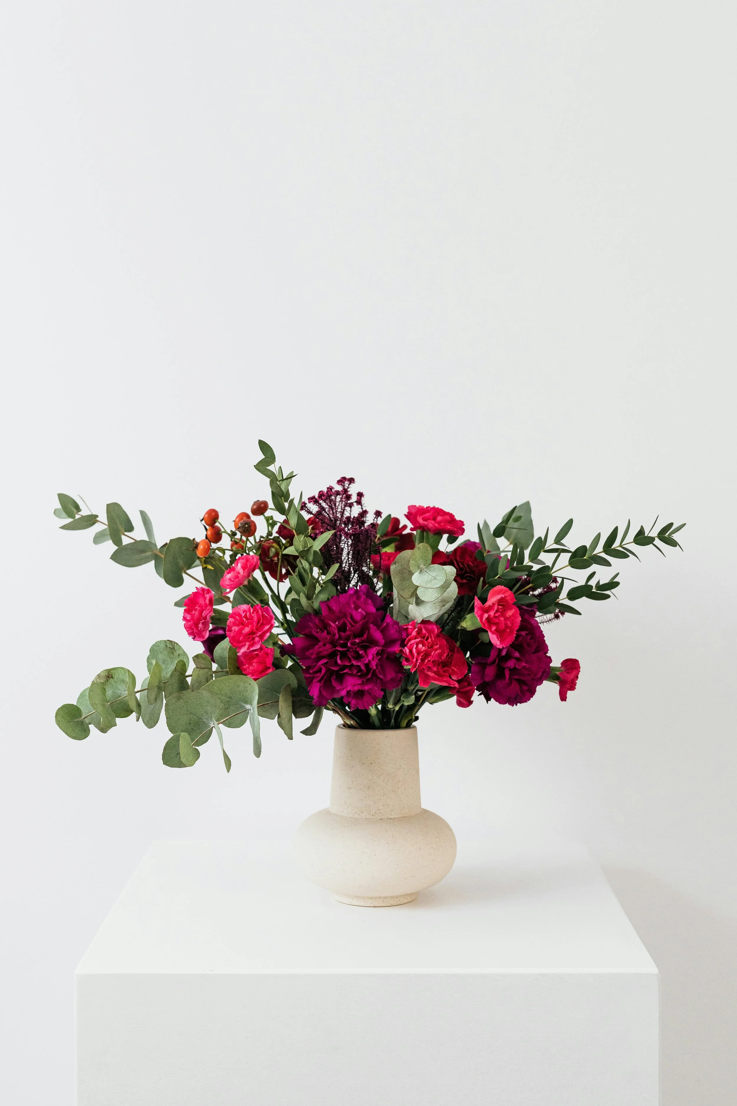 A beige vase with a bouquet of pink and burgundy flowers, green leaves, and berries, displayed on a white pedestal against a plain white background.