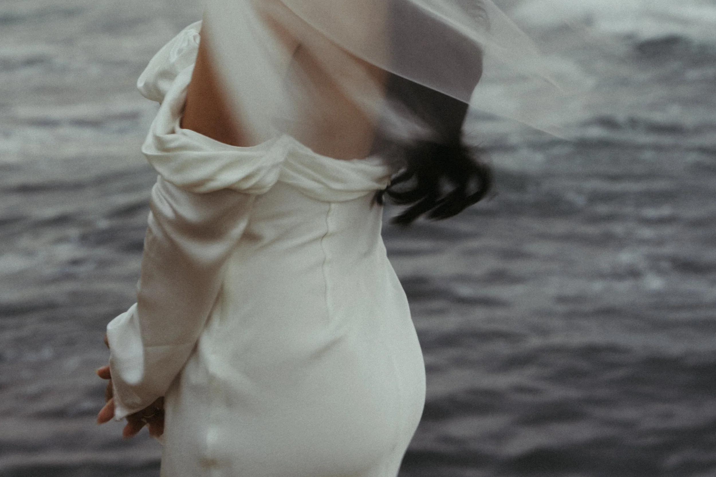 A woman in a white dress standing near a body of water, with her head turned away and her dark hair partially visible.
