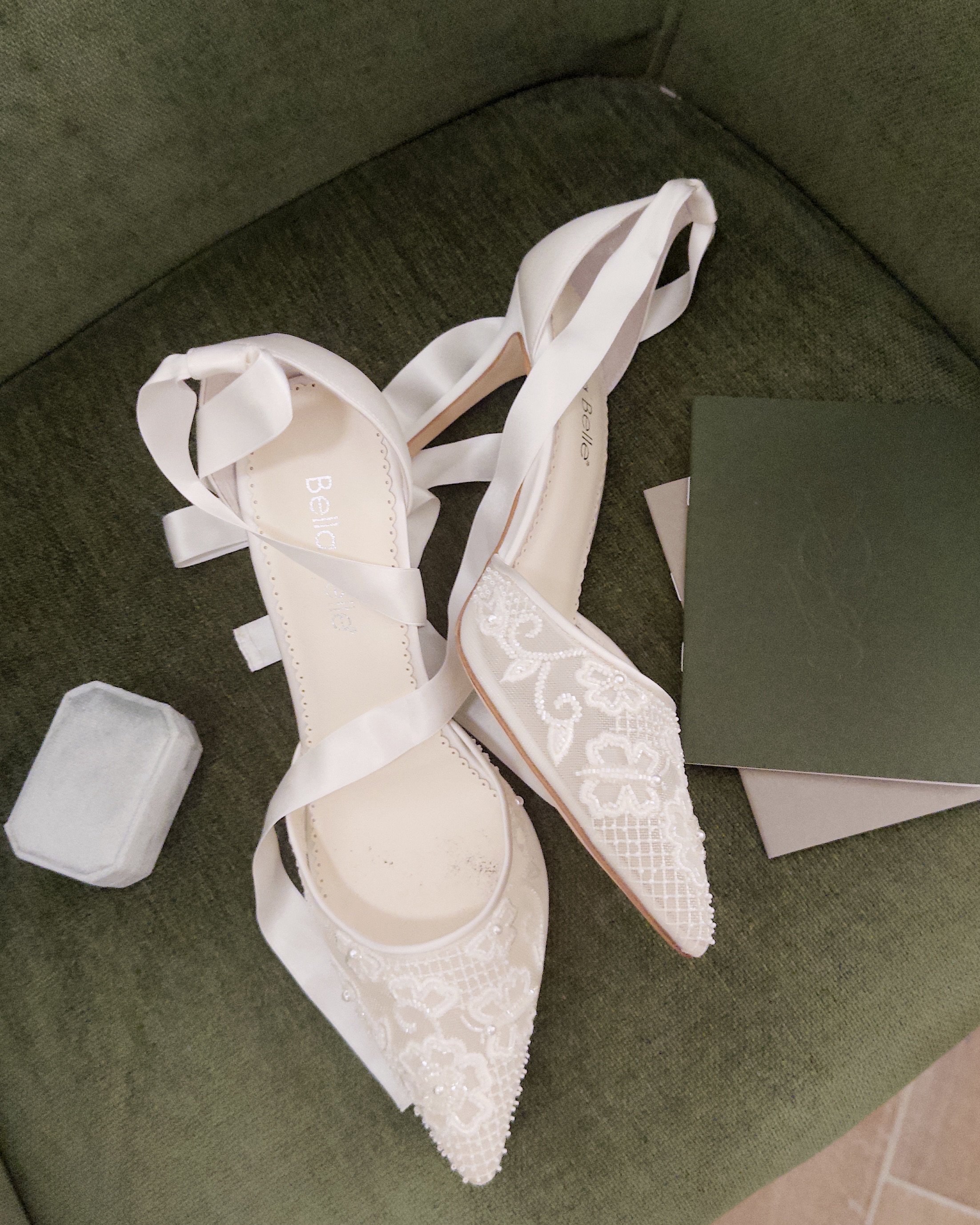 A pair of white bridal shoes with embroidered lace detailing and ribbon ties, resting on a green cushion, next to a small transparent box and an open dark-colored gift box.