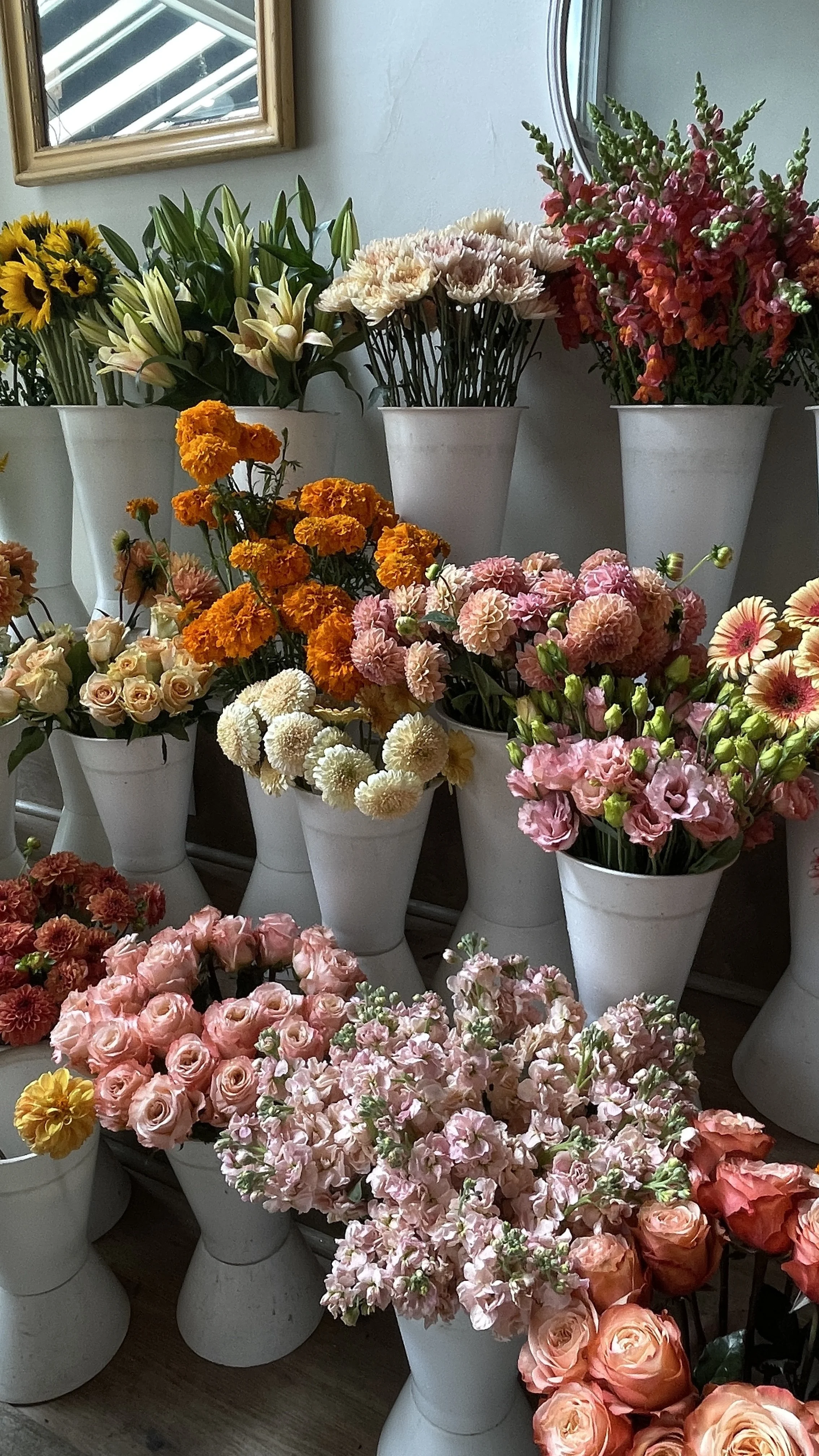Various colorful flowers in white vases on a wooden surface near a wall and window.