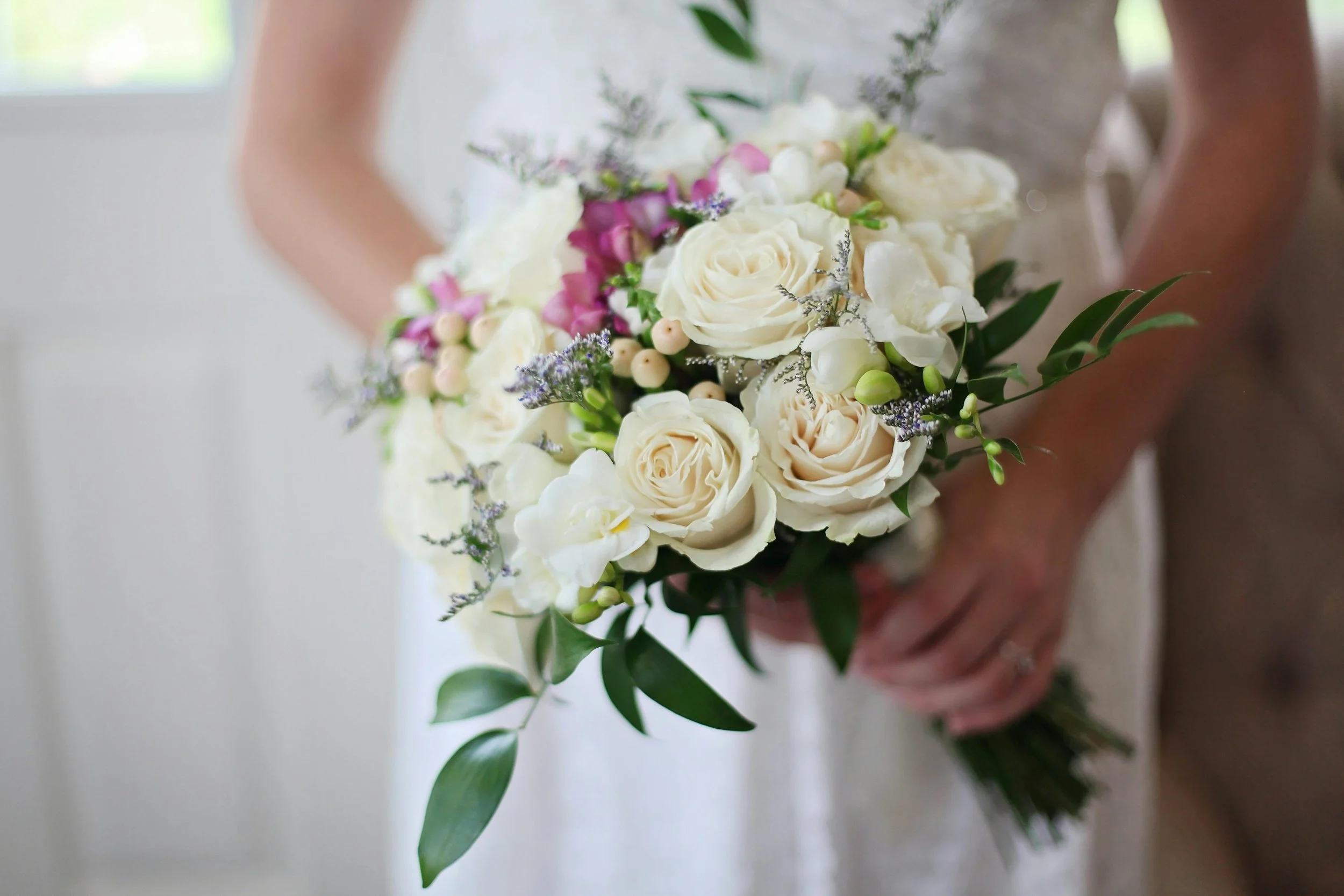 A bride holding a bouquet of white roses, cream-colored orchids, and purple accents with green leaves.