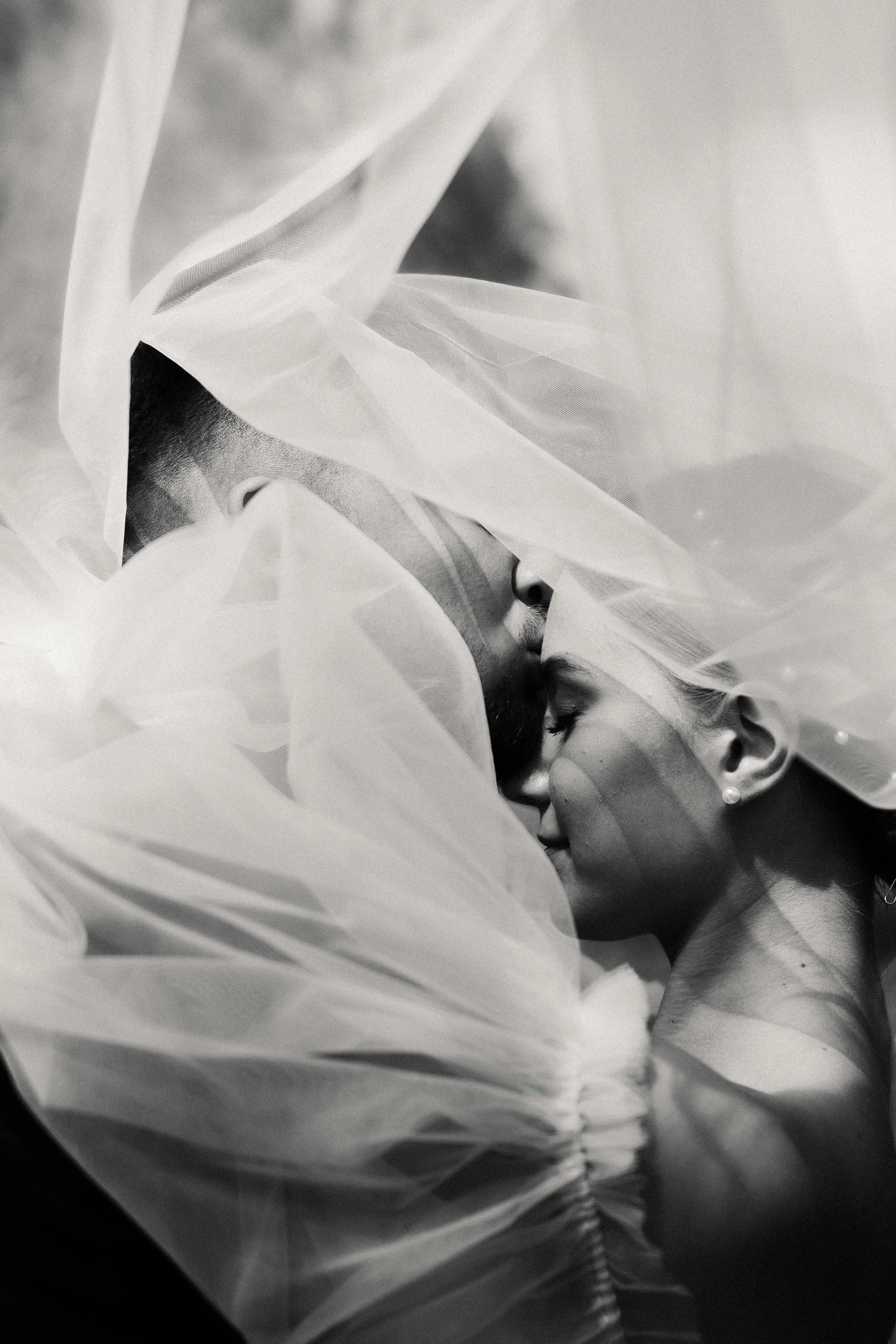 A black and white photo of a man and woman kissing, with sheer fabric partially covering their faces and shoulders.
