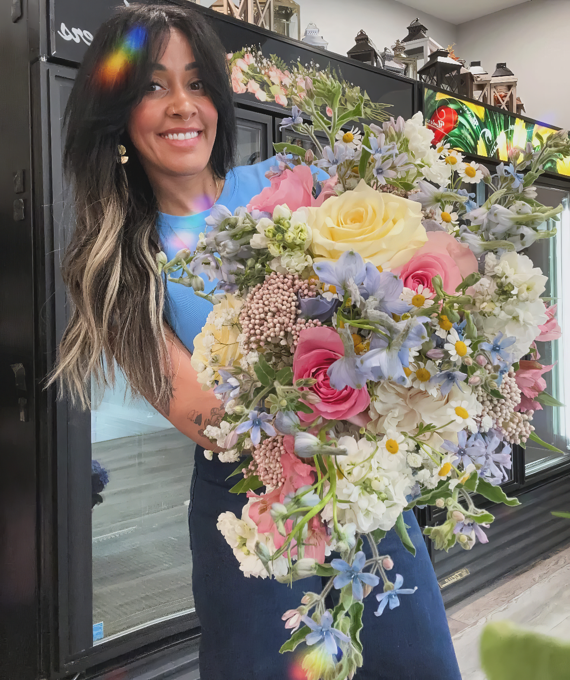 A woman with long dark hair holding a large, colorful bouquet of flowers, smiling indoors.