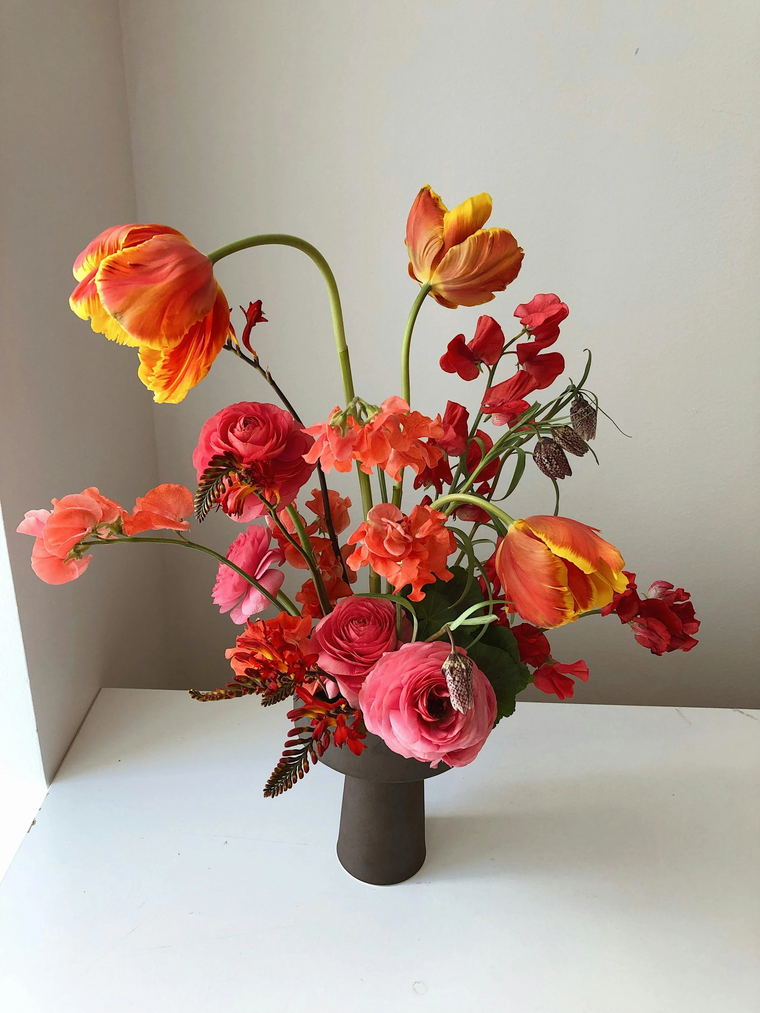 Colorful bouquet of pink, red, orange, and yellow flowers in a black vase on a white surface against a beige wall.