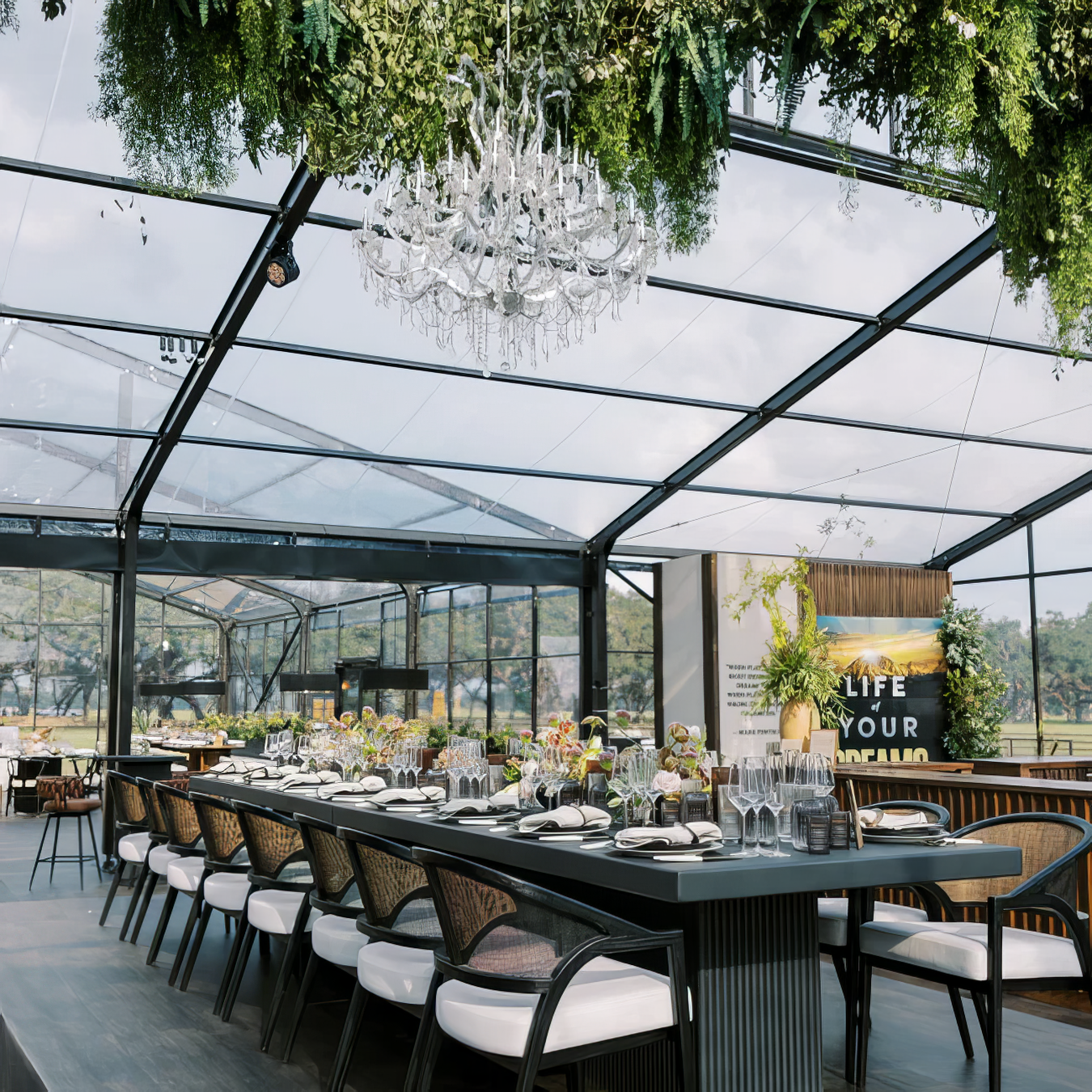 Elegant indoor event space with a long dining table set for a meal, decorated with flowers and glassware, under a glass ceiling with hanging greenery and a chandelier.