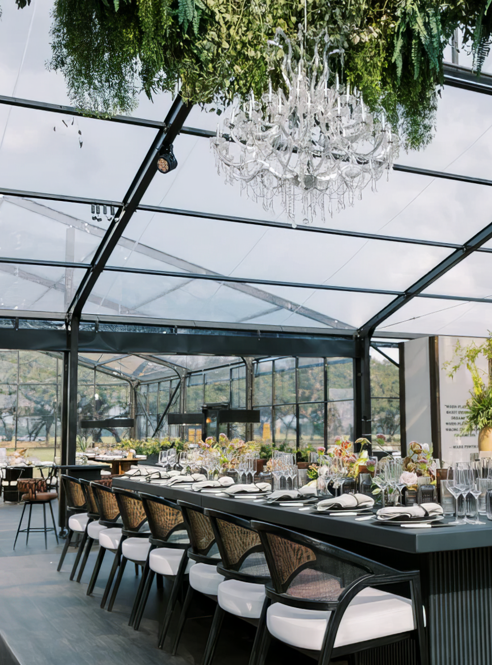 Elegant indoor dining area with a glass ceiling, decorated with a large crystal chandelier and greenery hanging above a long table set for a meal with white napkins, glassware, and floral centerpieces.