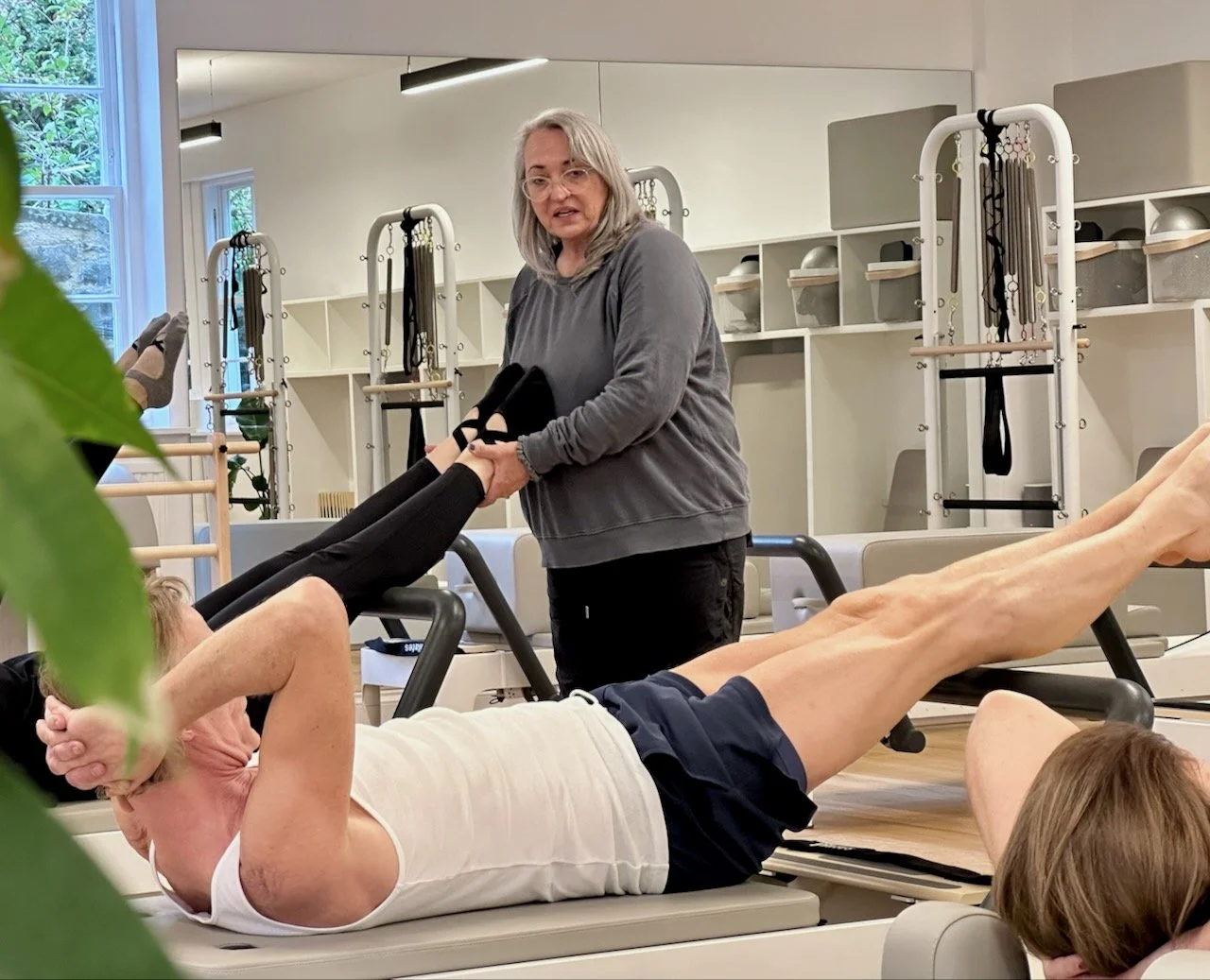Anna Marchington standing at a Reformer at Luma Pilates in Edinburgh, cueing a client's leg position during a class, with Reformers and the studio mirrored wall visible in the background.