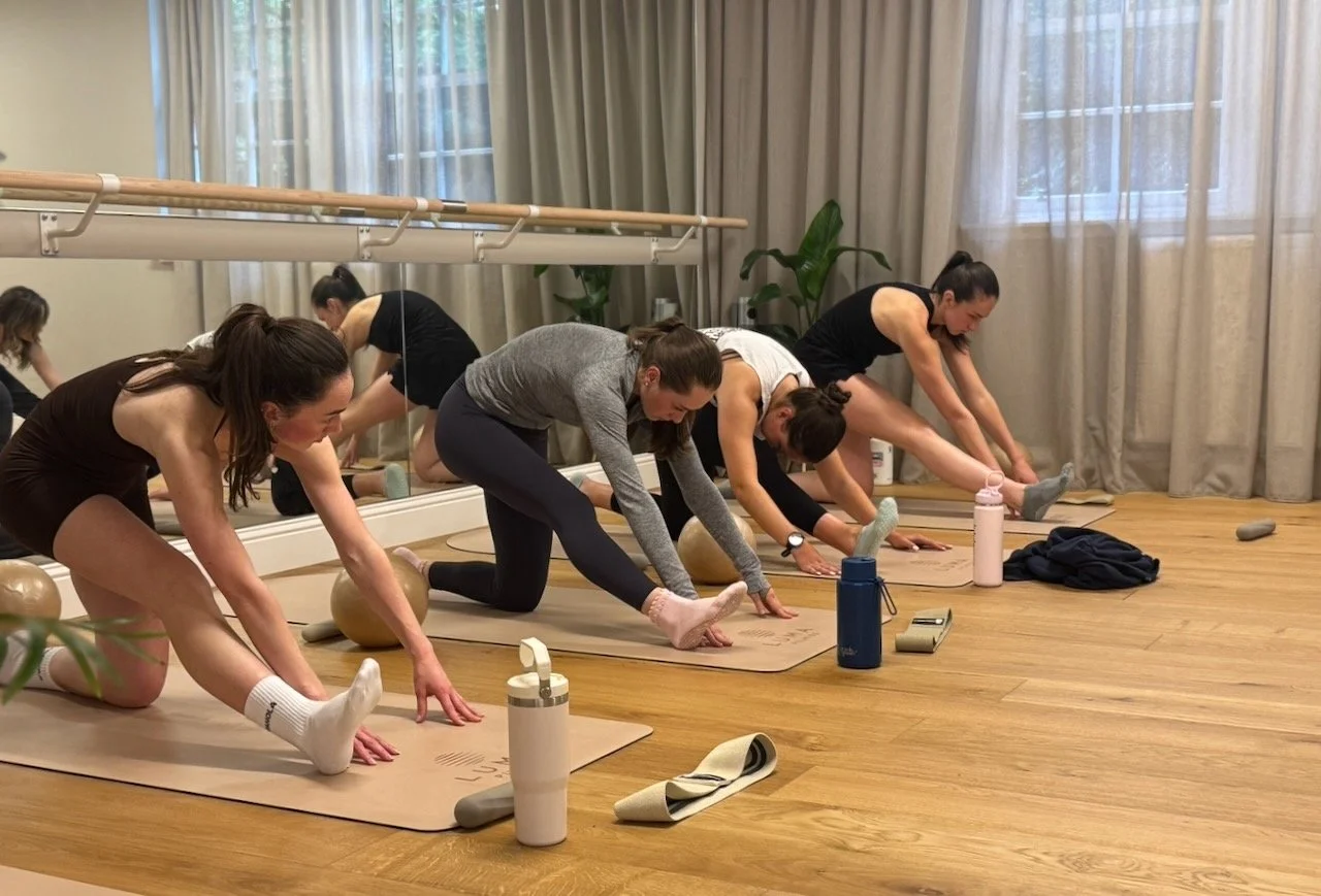 Clients stretching on mats at the end of a Barre class at Luma Pilates in Edinburgh, with the ballet barre and studio mirror visible behind them.