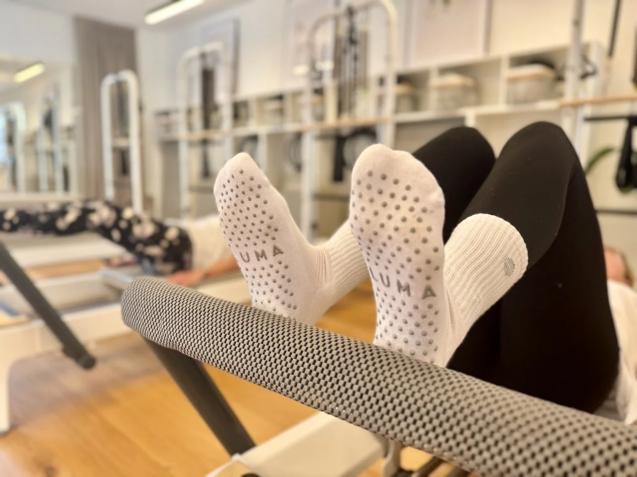 Close-up of Luma Pilates branded grip socks on a Reformer footbar during a class in Edinburgh, with clients and Reformers visible in the background