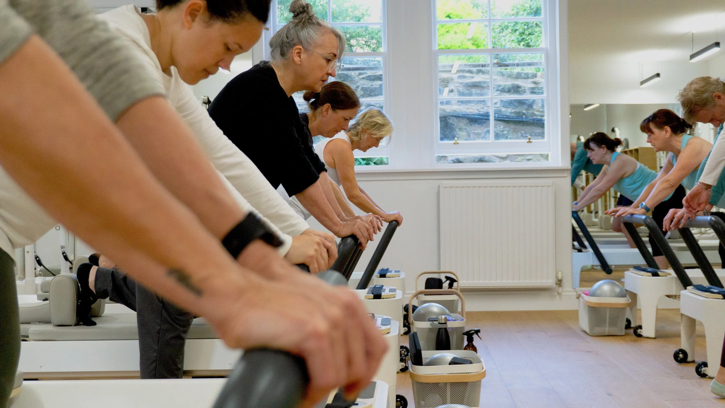 Group of diverse women in workout class, leaning forward on reformer machines, exercising in bright studio with large windows and wooden floors.