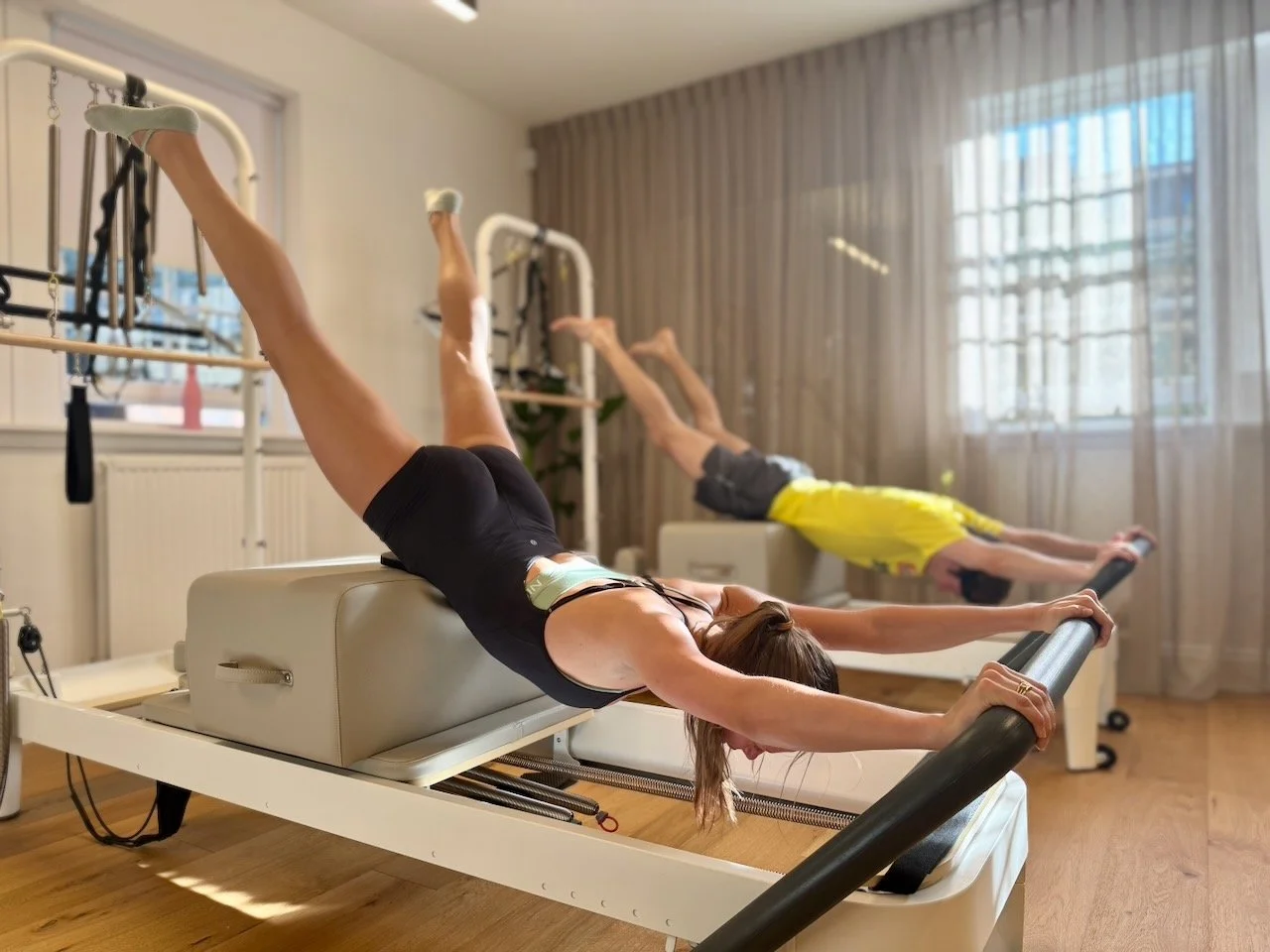 Two clients performing swan dive on the box on Reformers at Luma Pilates in Edinburgh, with the studio's New Town windows and natural light visible behind them.