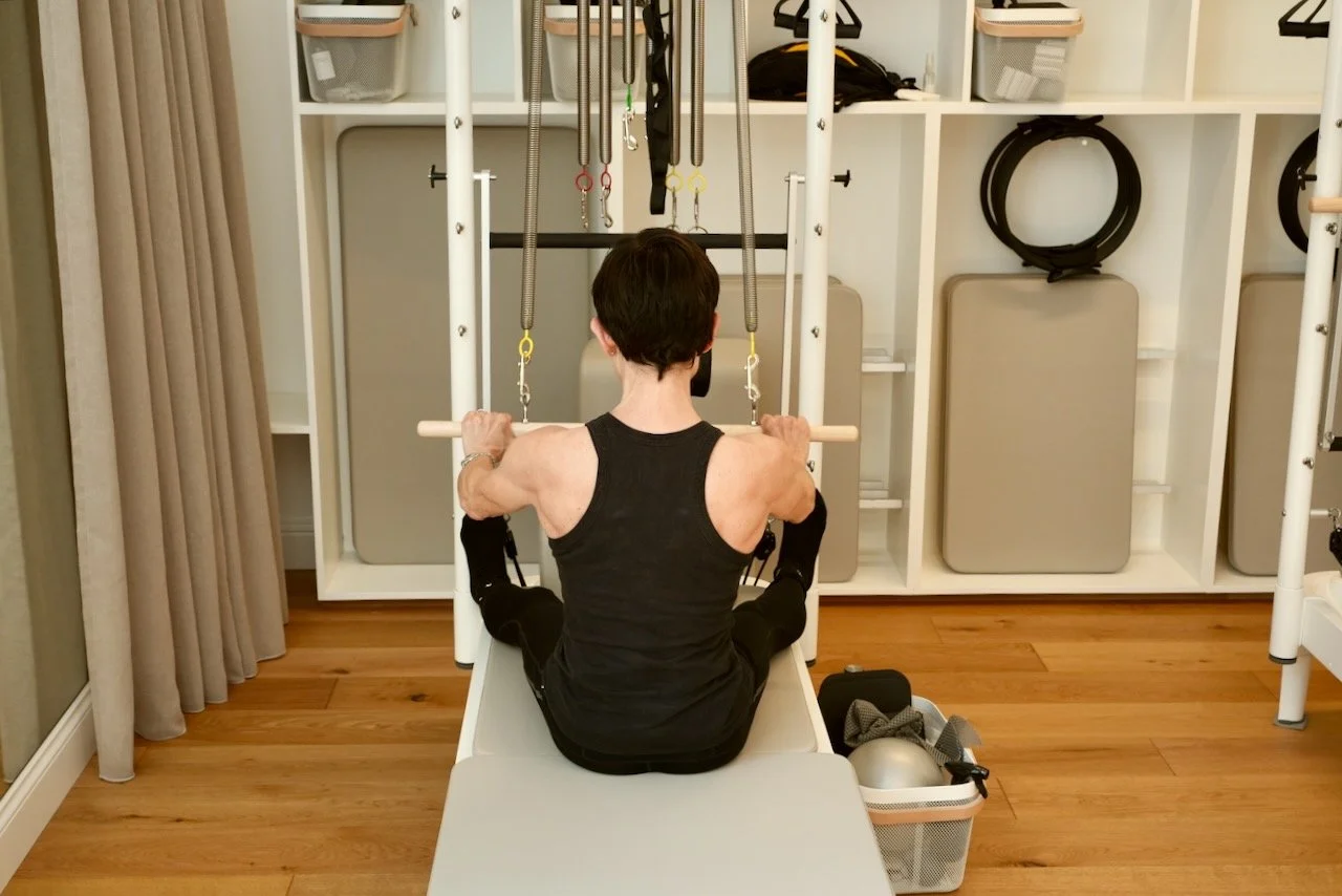 Client performing lat presses on the Tower at Luma Pilates Edinburgh, viewed from behind, with exceptional shoulder girdle alignment and the studio prop shelving visible in the background