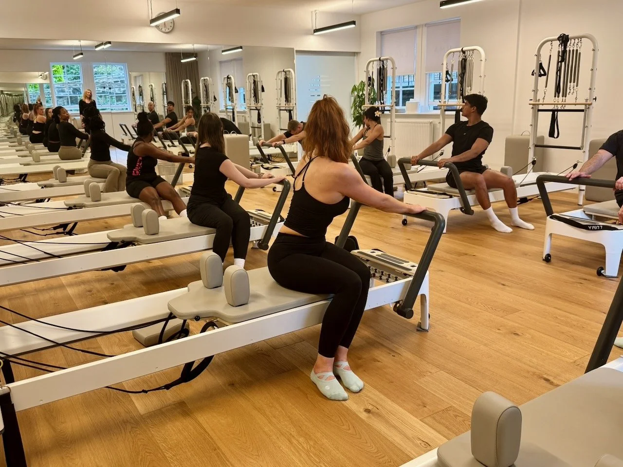 A full group Reformer class at Luma Pilates in Edinburgh performing a seated mermaid stretch and spine twist sequence, with Lucia Poulter visible at the front of the studio and the mirrored wall reflecting the full class behind.