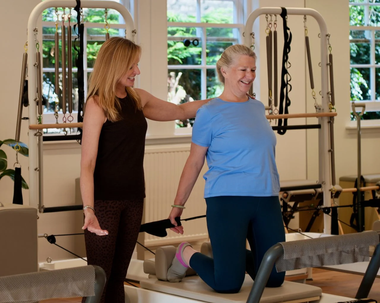 Lucia Poulter cueing a client to guide her alignment during a kneeling Reformer Pilates class. The client is smiling and holding a strap. A Tower frame is visible in the background.