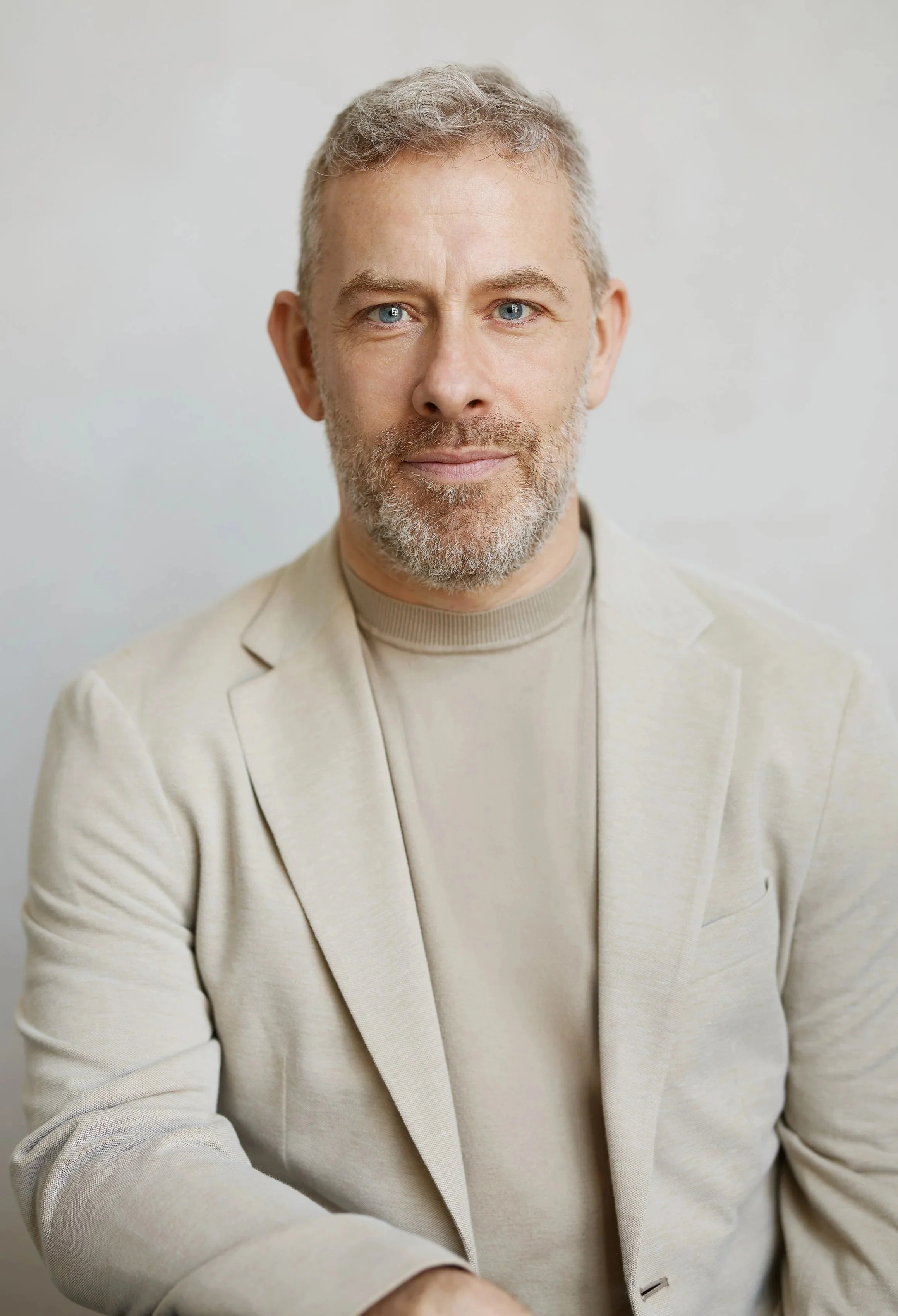 A middle-aged man with gray hair and a beard, dressed in a beige blazer and turtleneck, posing against a plain light background.