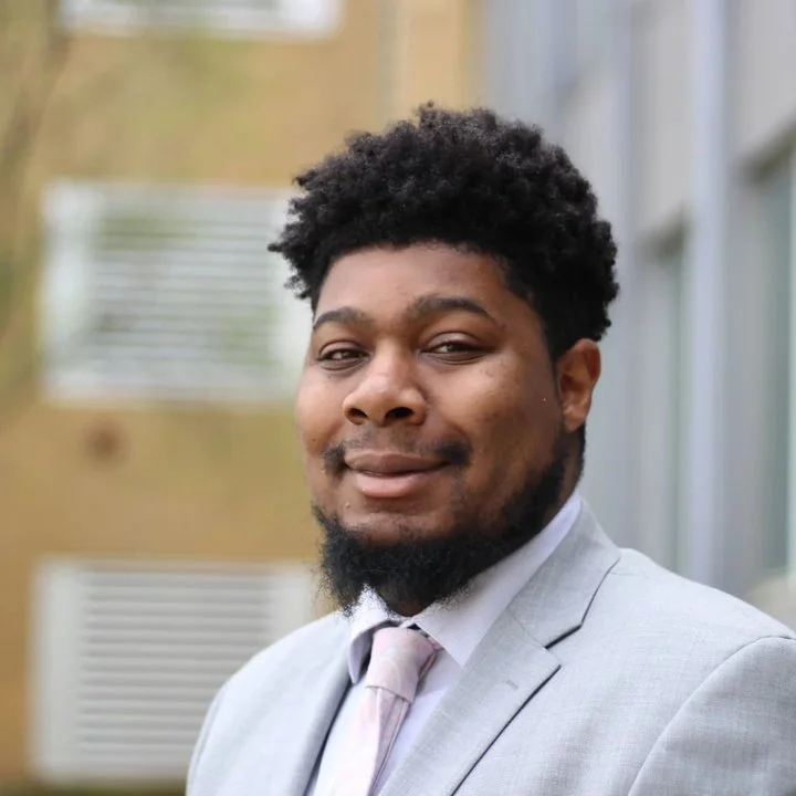 Portrait of an African-American or Black Attorney with dark curly hair and beard, wearing a light gray suit and pink tie, standing outdoors near a building with windows.